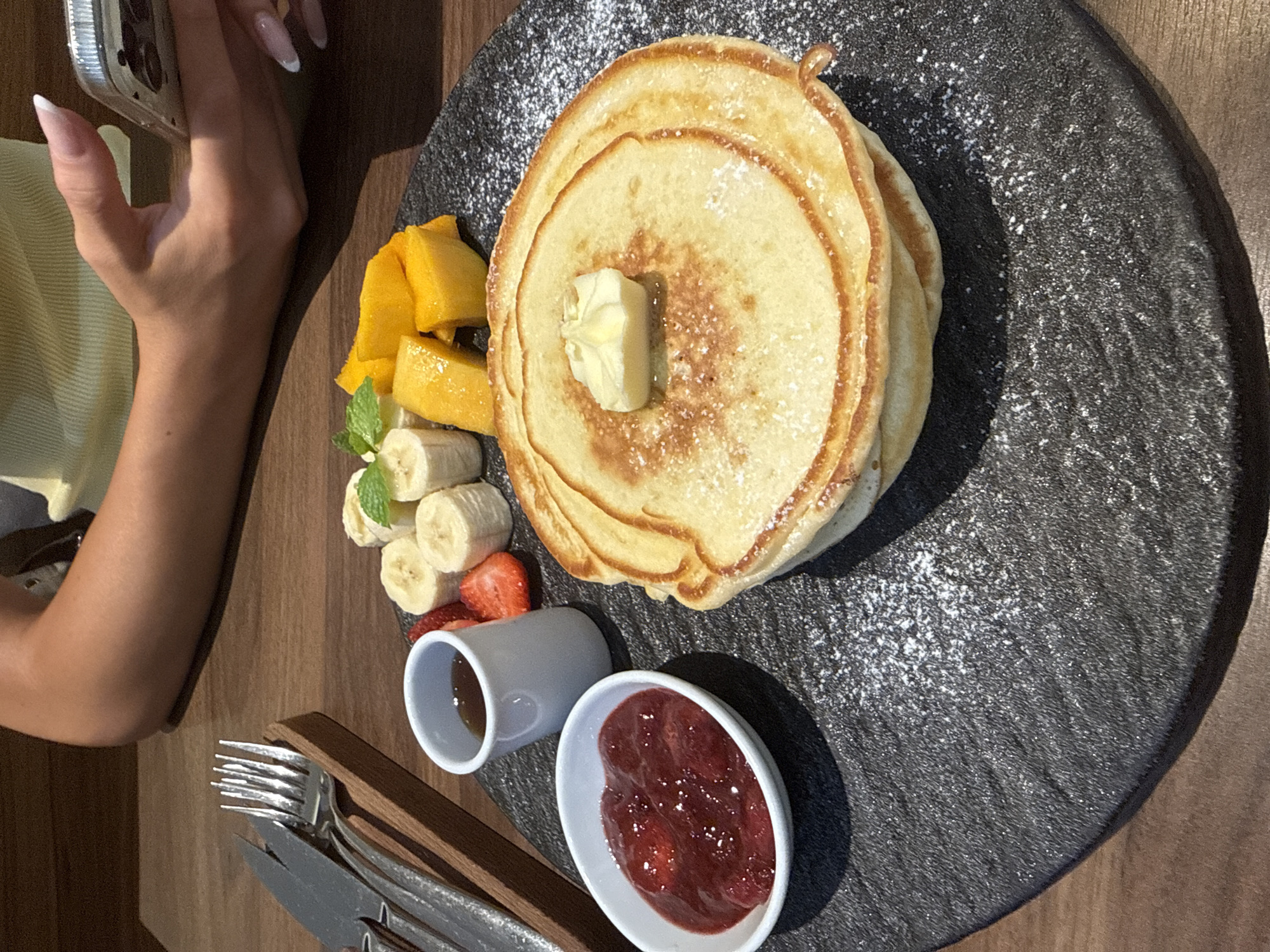 A high-angle shot captures a breakfast setting on a wooden table, likely in a café in Đà Nẵng, Vietnam. A woman's arm and hand are visible on the left, holding a phone. The main subject is a black textured plate featuring a stack of pancakes topped with butter and dusted with powdered sugar. Beside the pancakes are cubed mango, sliced bananas garnished with mint, and a sliced strawberry. Accompanying the pancakes are two small white dishes: one contains a dark syrup, and the other a red strawberry jam. Silverware, consisting of a knife and fork, is visible near the bottom left edge. The table is a light brown wood.