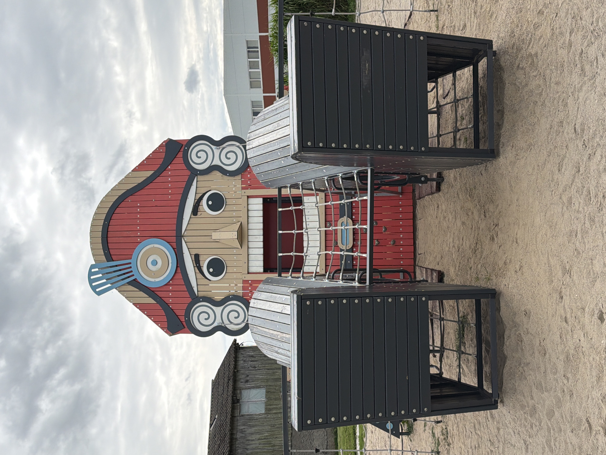 The image depicts a whimsical nutcracker-themed playground structure situated on a sandy surface. The nutcracker's face is rendered in wood, with large circular eyes, a prominent nose, and a wide, red-slatted mouth. A tall, red, wooden hat with a blue plume adorns its head. Flanking the nutcracker's face are two large, dark gray, wooden seating areas, each with a slatted backrest. A net-like rope structure is visible within the opening of the nutcracker, suggesting a climbing or entry point. In the background, to the left, is a rustic, wooden building with a pitched roof and a small window. To the right, a modern, white building with horizontal windows is visible. The sky is overcast with gray clouds. The scene is located in the city of Nekrasovo, Russia.