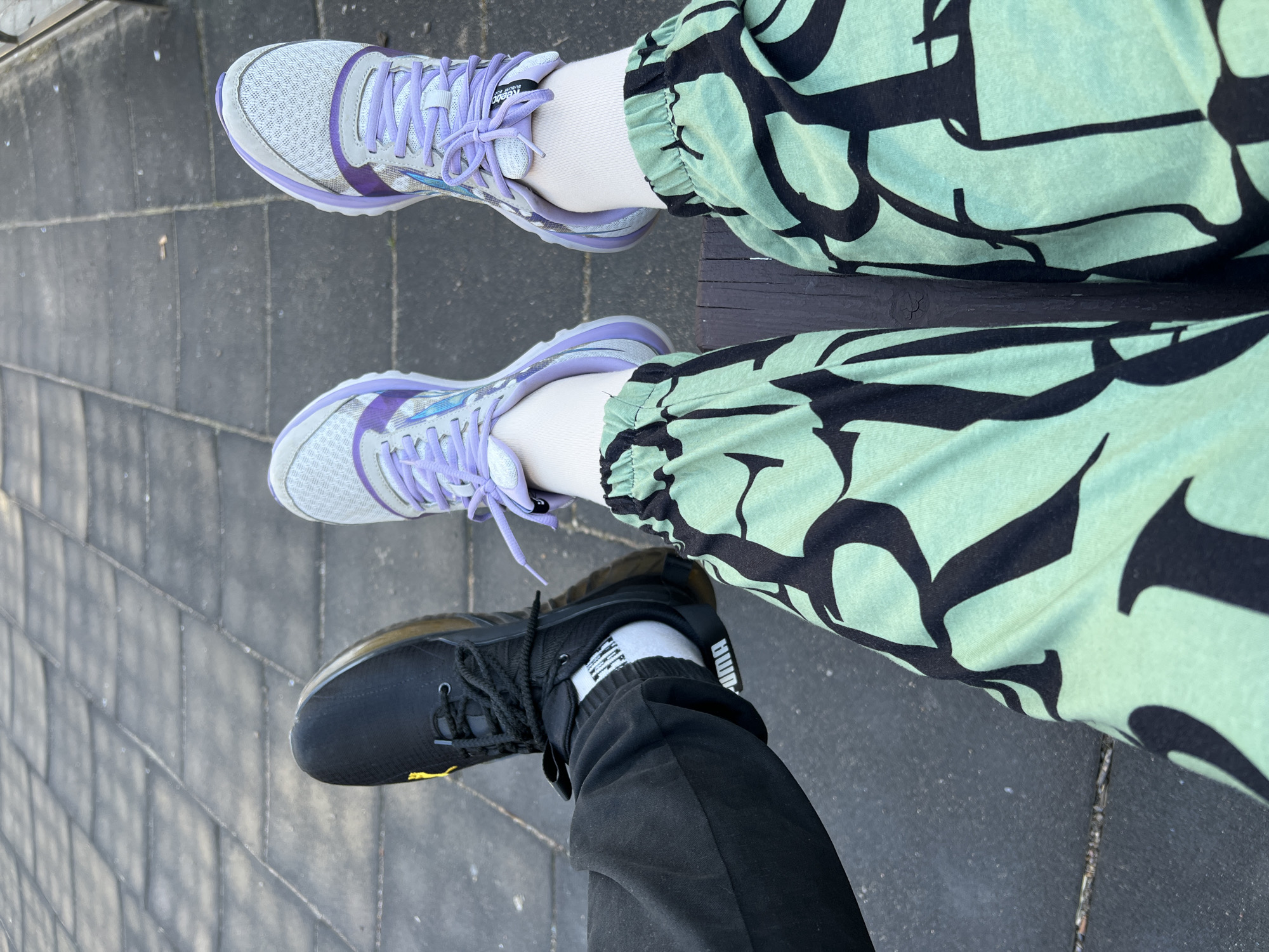 Two people's legs are shown from the knees down, seated on what appears to be a park bench or similar outdoor furniture. The ground beneath them is paved with dark, square-shaped tiles, typical of urban outdoor areas.
The person on the left is wearing black pants and black Puma sneakers with white socks. The person on the right is wearing light green pants with a black abstract pattern and white socks with light purple and white athletic shoes. The backdrop suggests an outdoor, possibly urban, setting. The image quality is clear, with good lighting. The location is identified as the city of Sinyavino, Russia.