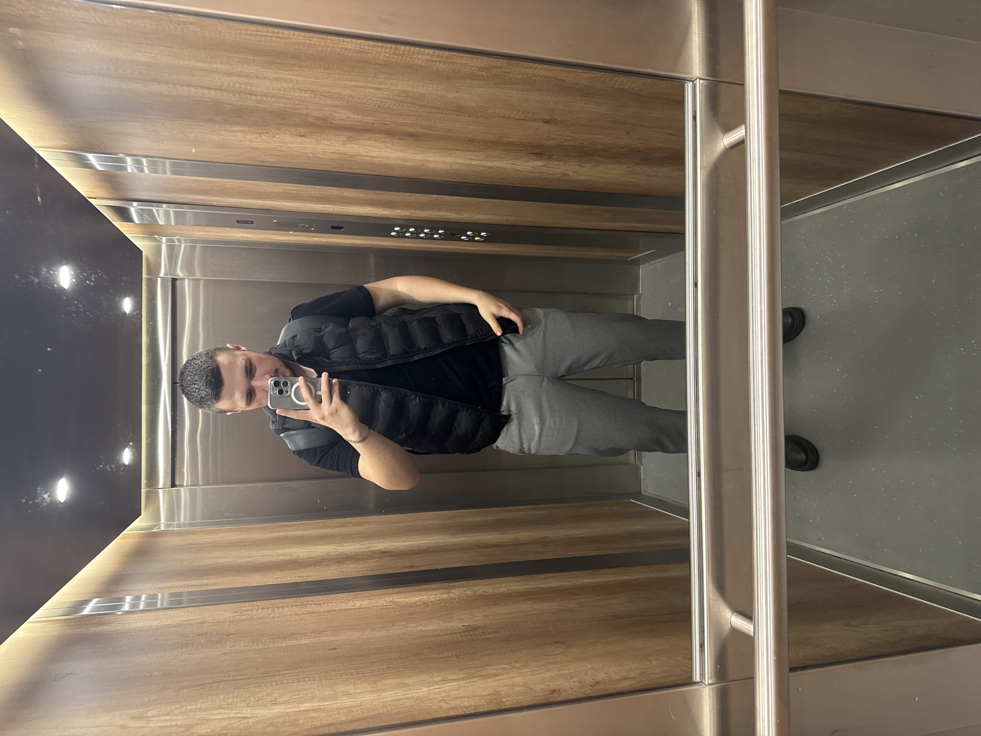 A person is standing in an elevator, taking a selfie. The elevator has wood-paneled walls with stainless steel accents and a gray floor. The elevator's floor indicator panel is visible near the top. The person is wearing a black t-shirt, a black vest, and gray sweatpants. There is no specific information to determine the location in Sofia, Bulgaria.