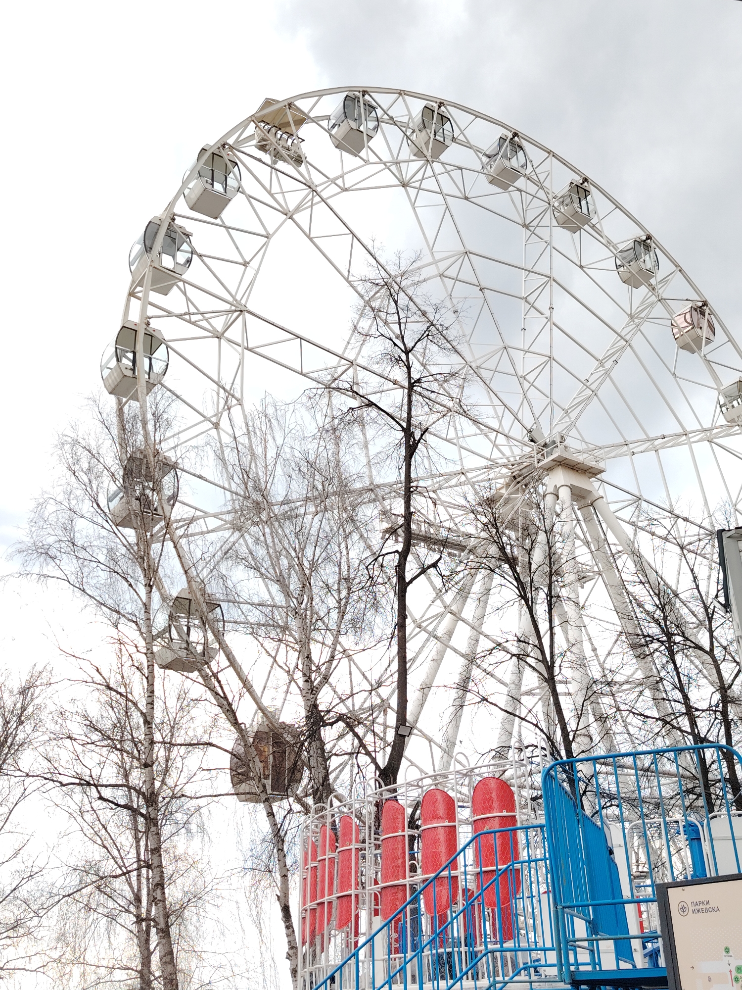 The image shows a Ferris wheel, likely in a park in Izhevsk, Russia, based on the park information sign. The Ferris wheel has white structural supports and enclosed cabins. Several leafless trees partially obstruct the view of the Ferris wheel. A blue metal fence and red padded pillars are in the foreground, possibly part of the entrance to the Ferris wheel or a nearby ride. The sky is overcast. No people are visible in the frame.