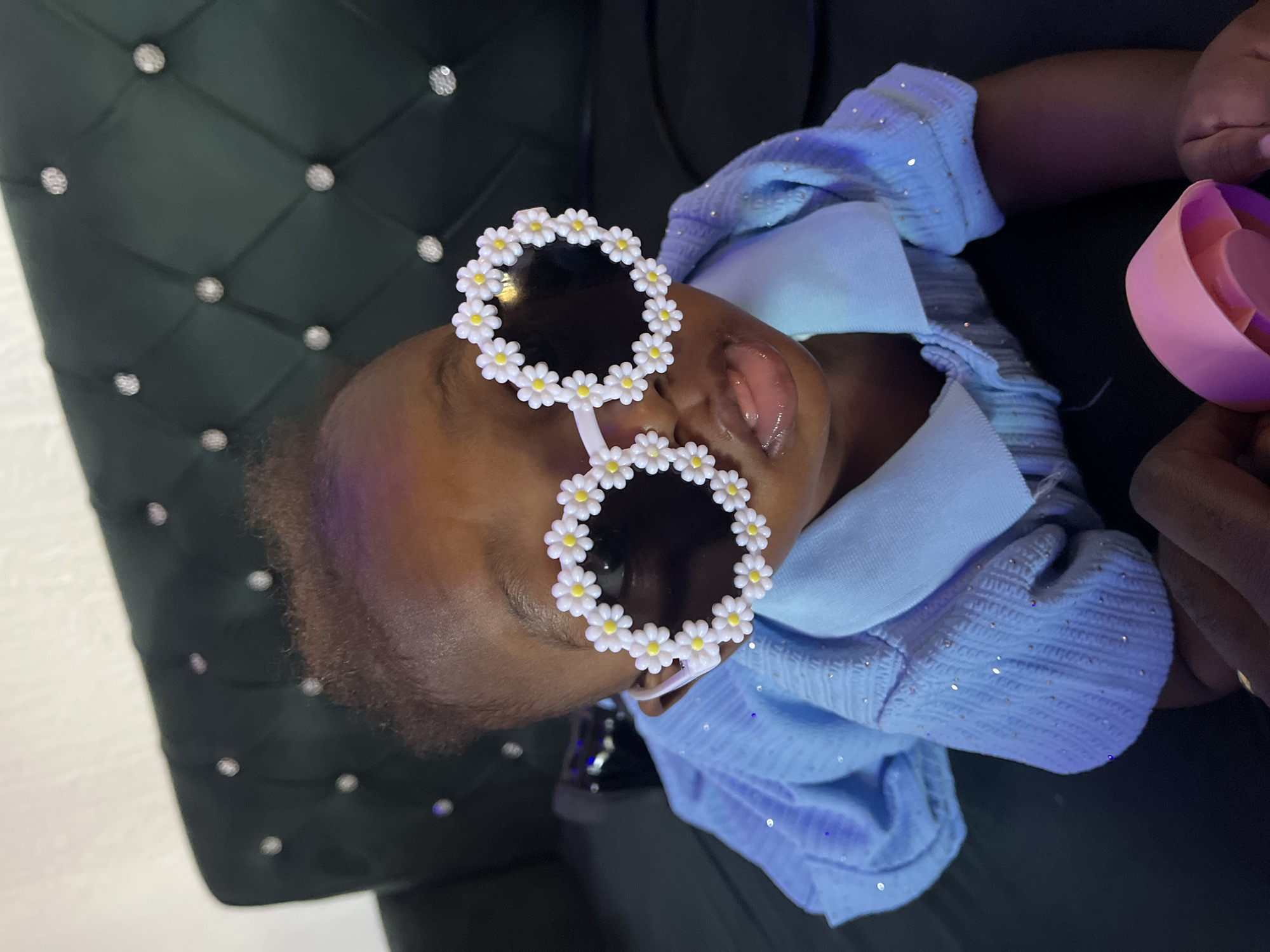 A close-up shot captures a young child lying on a dark, tufted surface that appears to be a sofa. The child is wearing a light blue, sparkly shirt with a collar and is looking upwards, slightly to the right. The child is also wearing oversized white sunglasses adorned with small white daisies, each with a yellow center. The child's tongue is slightly protruding. In the foreground, a person's hands are visible, holding a pink object. The lighting suggests an indoor setting. The image does not provide any visual cues or context to determine a specific location within Barakin Akawo, Nigeria.