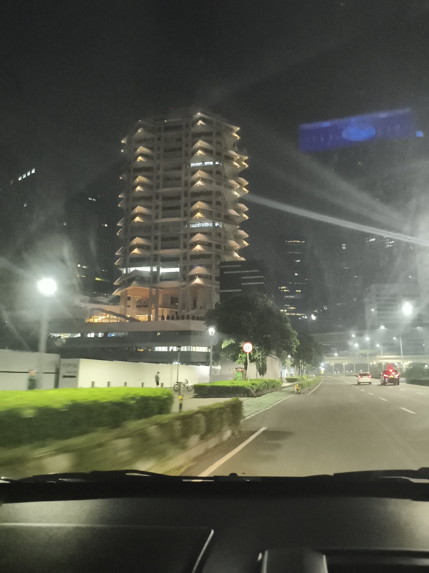 The image depicts a nighttime scene in South Jakarta, Indonesia, captured from the interior of a moving vehicle. The roadway stretches into the distance, illuminated by streetlights. Two vehicles with illuminated taillights are visible ahead. A tall, tiered building with numerous illuminated balconies dominates the left side of the frame. There are trees and green bushes growing along the side of the road. A white wall is visible behind the bushes. A pedestrian and a parked motorbike are near the wall. The vehicle's dashboard and windshield are in the foreground. Light refracts on the windshield. Other buildings are faintly visible in the background.