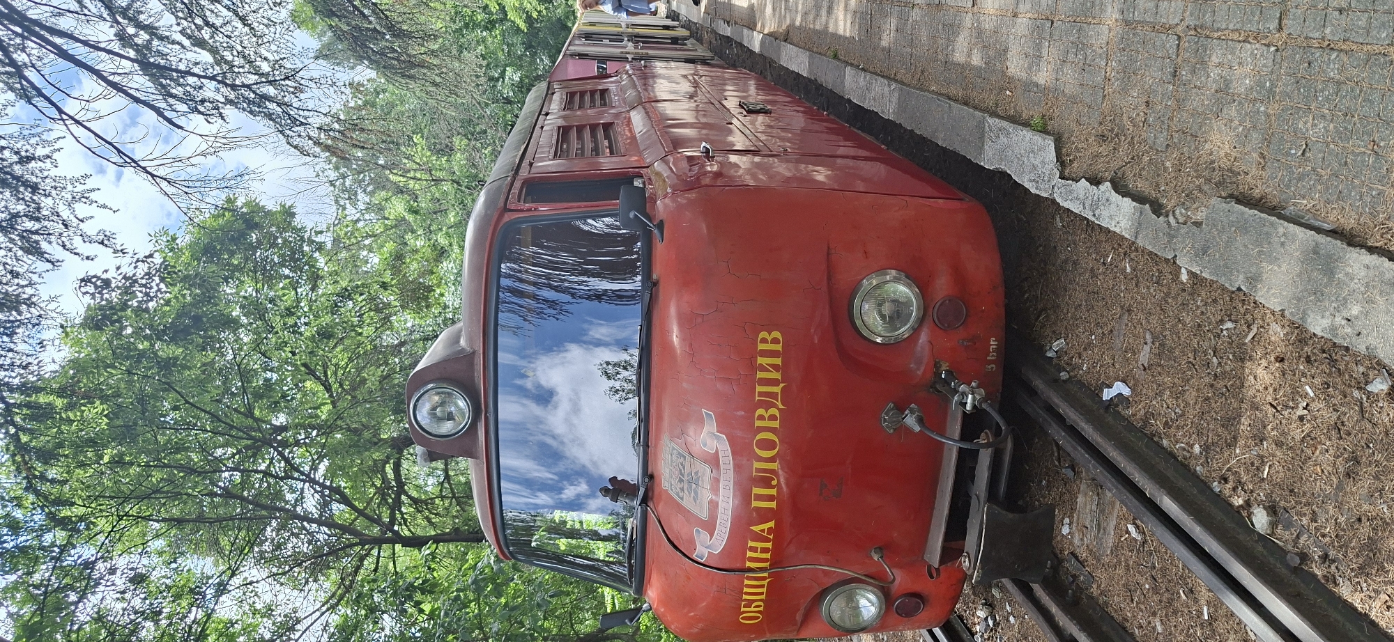 The image shows the front of a red vintage train car with yellow Cyrillic script reading "ОБЩИНА ПЛОВДИВ" ("Municipality of Plovdiv") on its front panel. The train is situated on a narrow-gauge railway track flanked by greenery and trees. The sky is visible through the train's windshield. The setting appears to be a park or recreational area, with the train potentially part of a children's railway or a historical exhibit. The train car exhibits signs of wear and tear, with visible cracks and aged paint. A concrete walkway or curb is adjacent to the railway track, with fallen leaves and debris scattered on the ground.