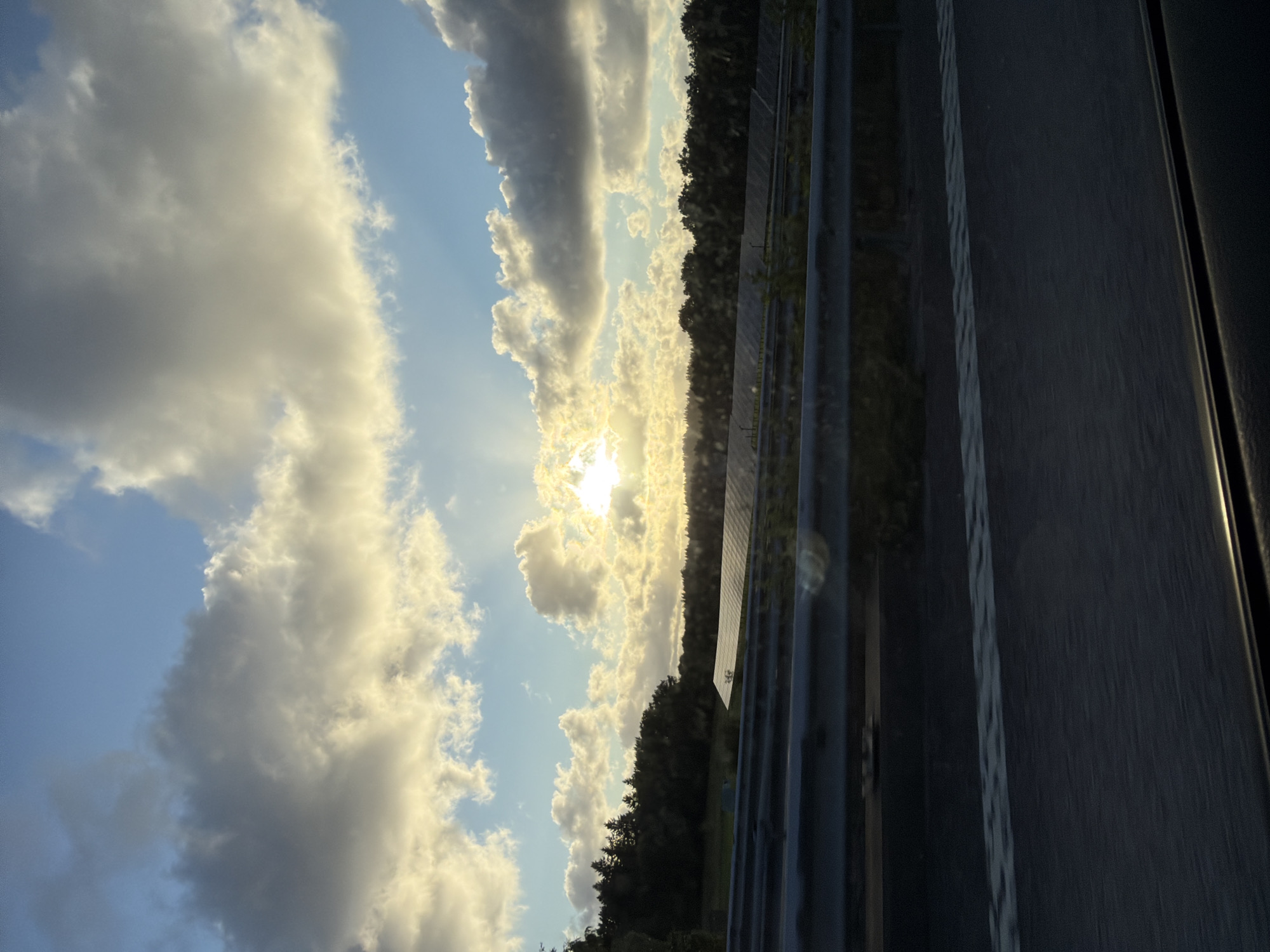 The image depicts a solar panel farm situated in Ödsmål, Sweden, during sunset. The foreground shows a highway barrier, indicating the viewer is likely traveling by car. Beyond the barrier, rows of solar panels are visible, stretching across a field backed by a dense line of trees. The sky is filled with dramatic clouds illuminated by the setting sun, casting a warm glow over the landscape. The overall scene suggests a rural or semi-rural environment in Sweden.