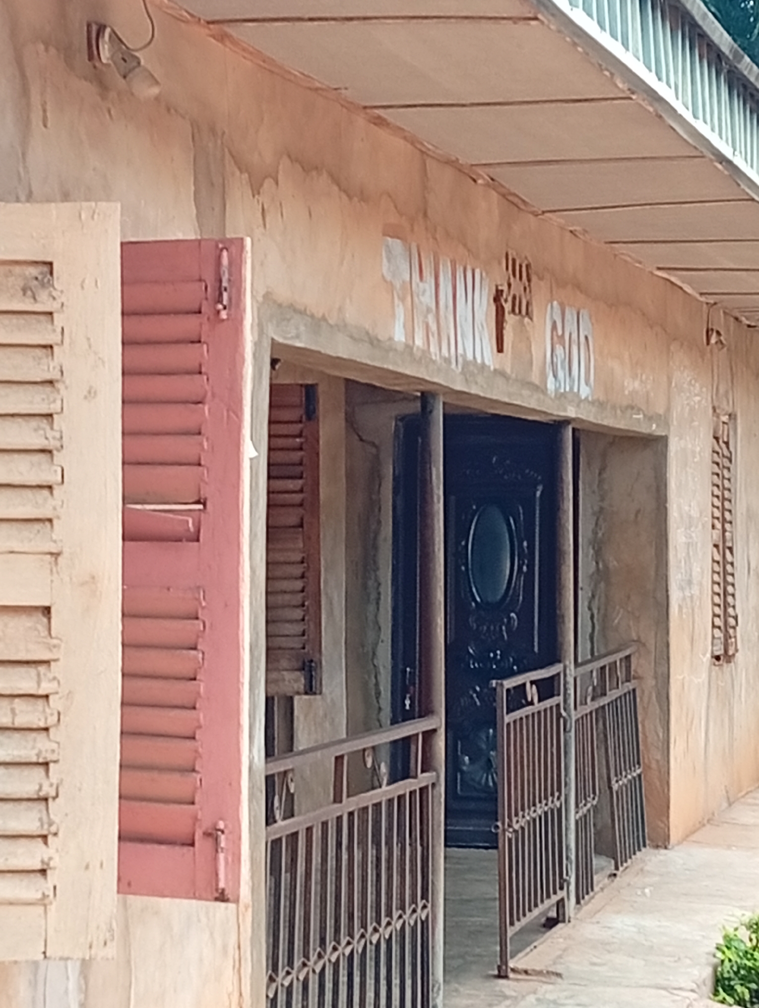 The image depicts the exterior of a building, likely a church or a community center, in Adda, Nigeria. The building's facade is rendered in a light, weathered plaster. Above the entrance, the words "THANK GOD" are painted in white, with a stylized depiction of a raised hand or cross between "THANK" and "GOD". The entrance features a dark, ornate double door with an oval window. Flanking the entrance are two windows with red shutters, one of which is partially open, revealing slatted blinds. A metal fence with vertical bars and decorative diamond patterns surrounds the entrance area. To the right of the entrance, another window with similar slatted blinds is visible on the exterior wall. The ground in front of the building appears to be a concrete walkway. A single light fixture is mounted on the wall above and to the left of the entrance. The overall impression is one of a modest, functional building.