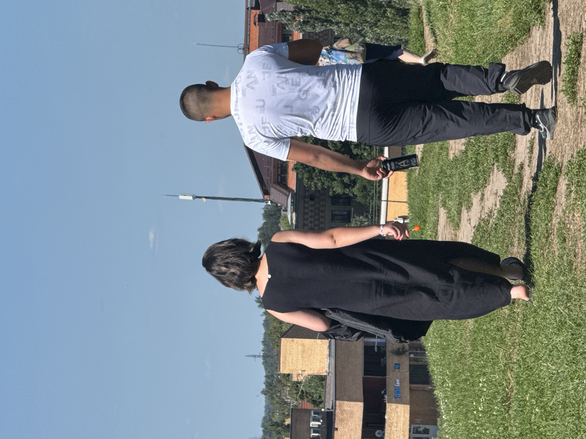 In an urban park setting in Dmitrov, Russia, a man and a woman walk away from the camera on a dirt path. The woman, on the left, wears a black sleeveless dress and appears to be barefoot, carrying a black bag over her left shoulder. She is also holding a small orange object in her right hand. The man, on the right, wears a white t-shirt with a repeating pattern of letters and black athletic pants. He holds a black can in his right hand. The path is bordered by green grass with small white flowers. In the background, there are several buildings with brick and beige facades, as well as trees and a clear blue sky with a few wispy clouds. Two cell towers are visible in the distance.