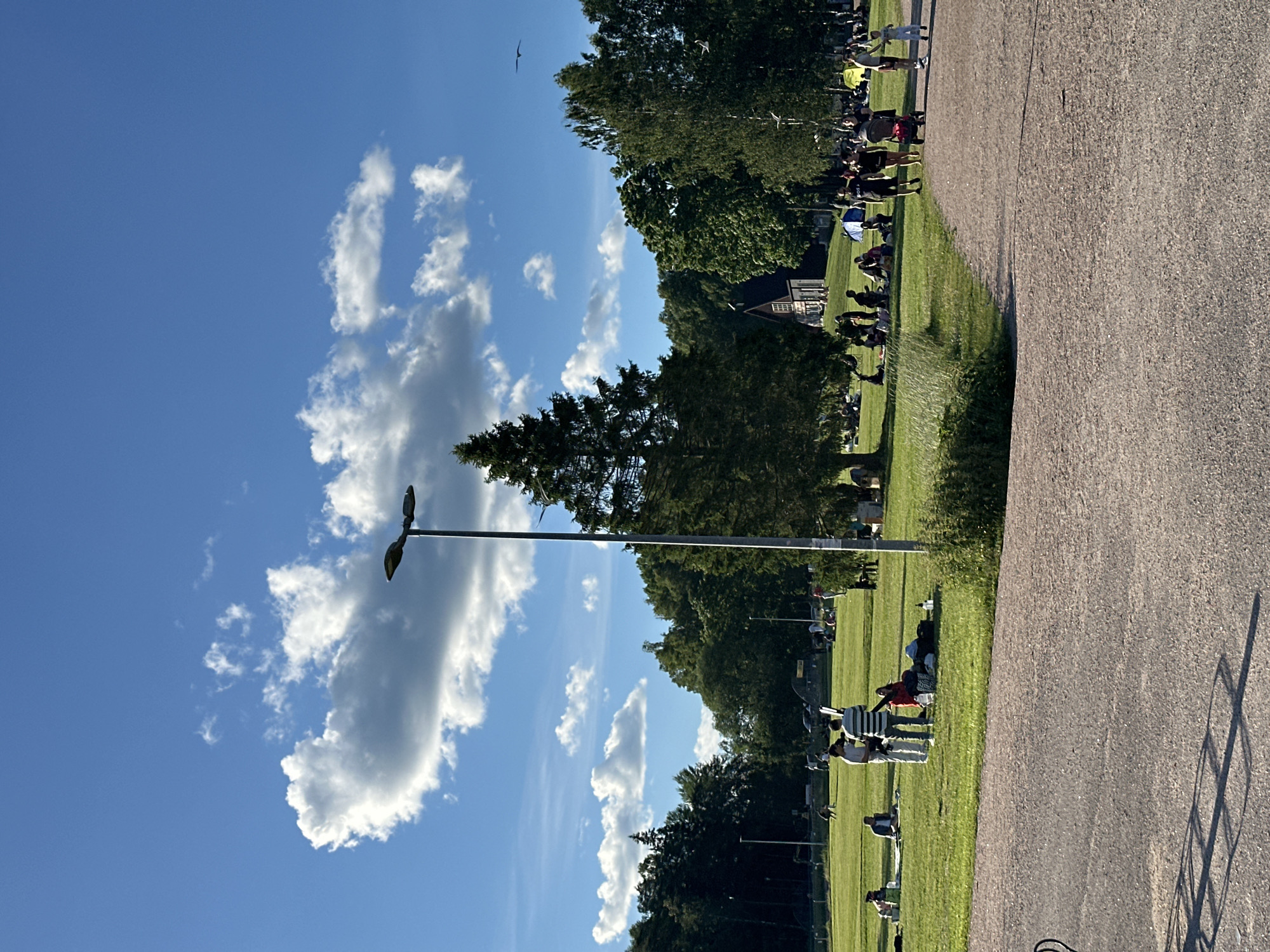 An outdoor scene depicts a park on a clear day with scattered clouds. A paved path runs through the foreground, with a grassy area beyond. Several groups of people are scattered across the grass, some sitting on blankets and others standing. A tall, thin streetlight stands in the middle ground. A small, dark building with a triangular roof is visible in the background, surrounded by trees. A bird is flying in the sky.