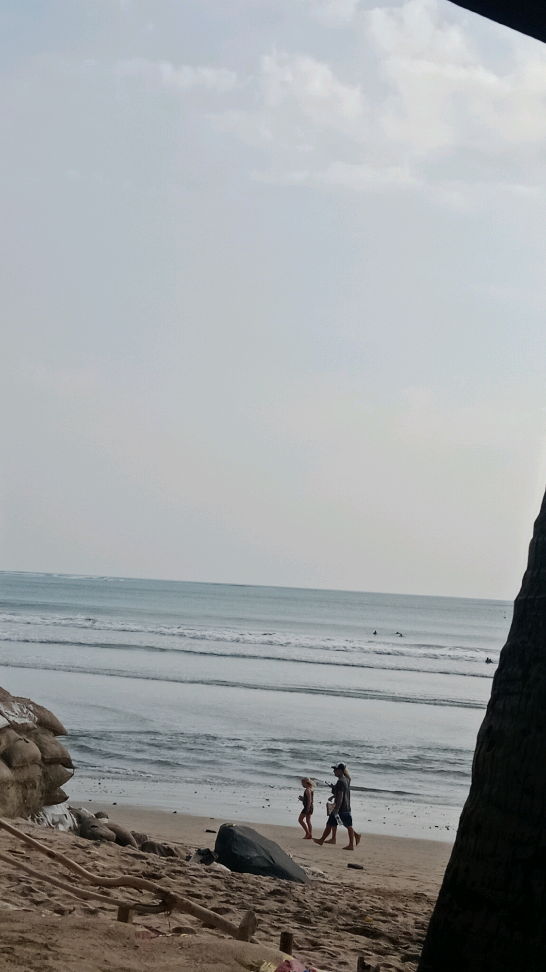 The image depicts a beach scene in Kuta, Indonesia. In the foreground, sandy beach with driftwood is visible. A few people are walking along the shore. In the midground, the ocean with gentle waves is present, and surfers are visible in the distance. The sky is overcast. The overall lighting suggests it is daytime, likely late afternoon.