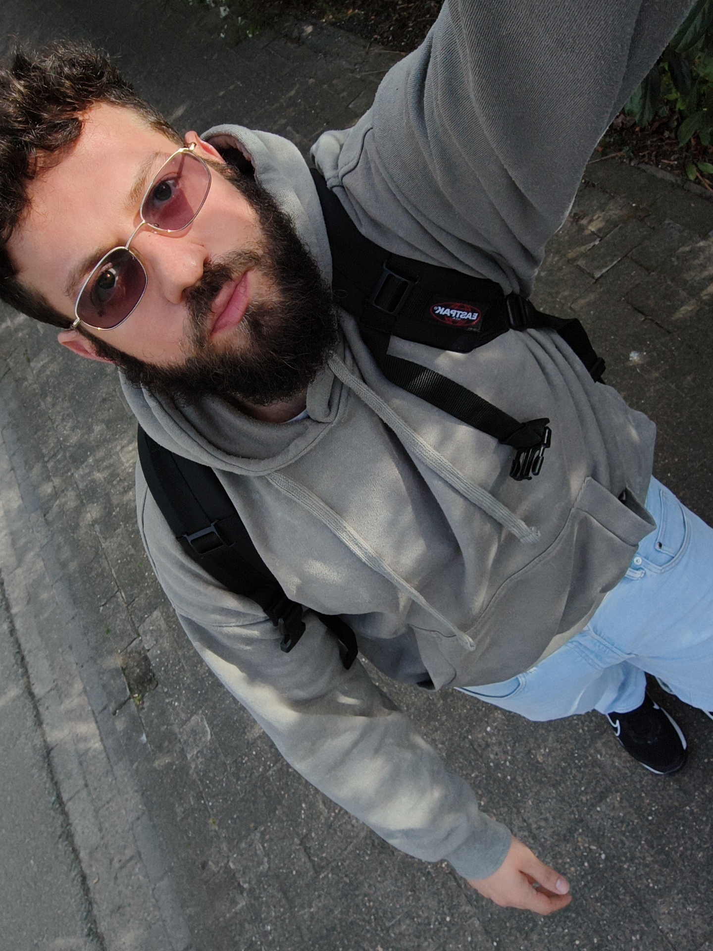 A man with a dark beard and sunglasses is standing on a gray concrete sidewalk. He is wearing a gray hooded sweatshirt and light blue jeans. He is wearing a black backpack with the word "EASTPAK" visible. A black and white shoe is visible on his right foot. The location is possibly a street in Diepholz, Germany, based on geographic proximity and typical European street aesthetics.