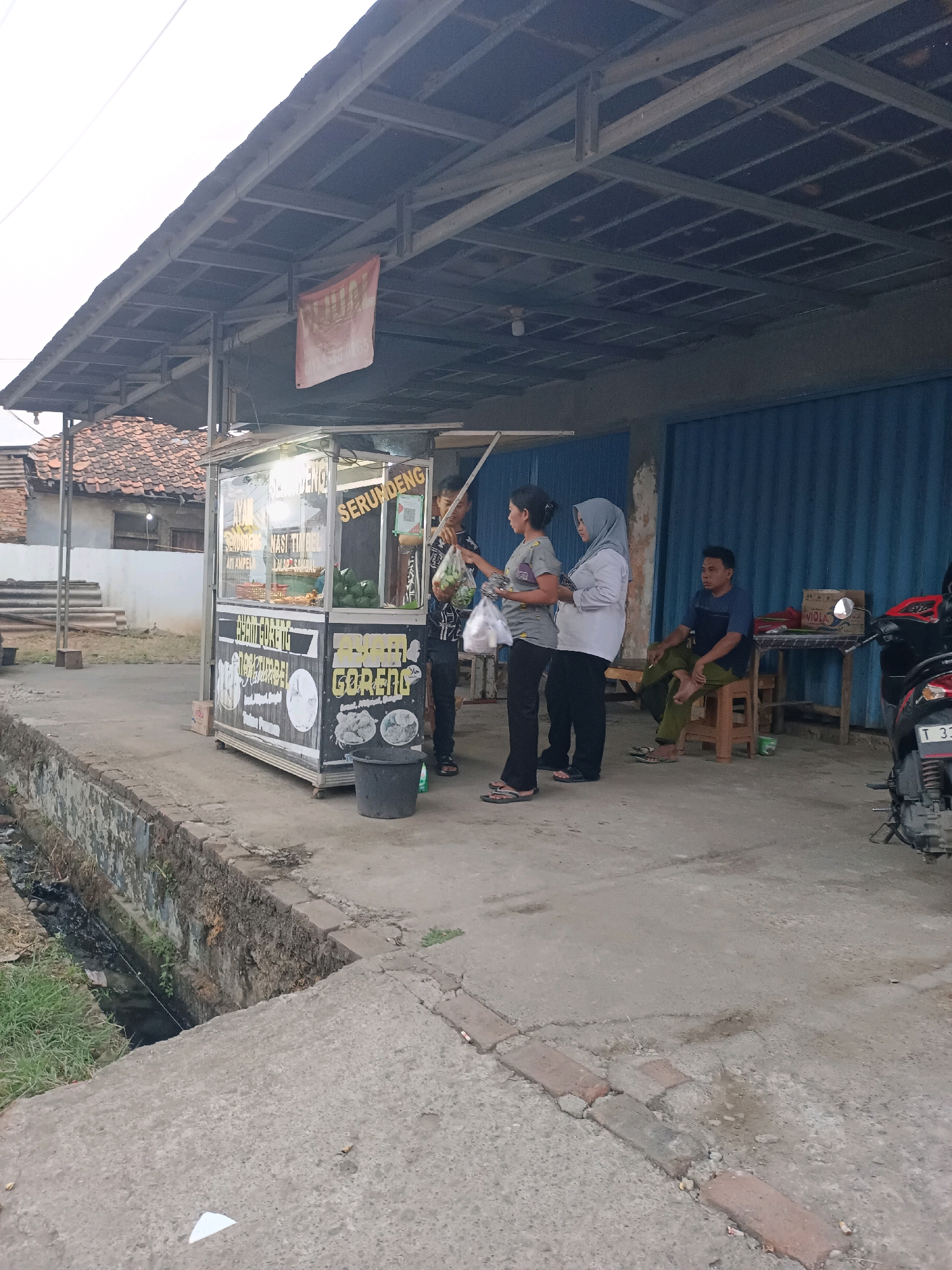 In the city of Karawang, Indonesia, an outdoor food stall is depicted. The stall, which appears to be a portable kiosk, displays a variety of food items and menu options. Several people are present: two women are standing at the counter, seemingly interacting with a vendor or making a purchase. Another man is seated on a stool to the right, observing the scene. In the background, a motorcycle is parked, suggesting the mode of transport for some individuals. The environment consists of a concrete walkway, a drainage ditch, and a building with a blue corrugated metal facade. The overall scene captures a moment of daily life at a local food vendor.