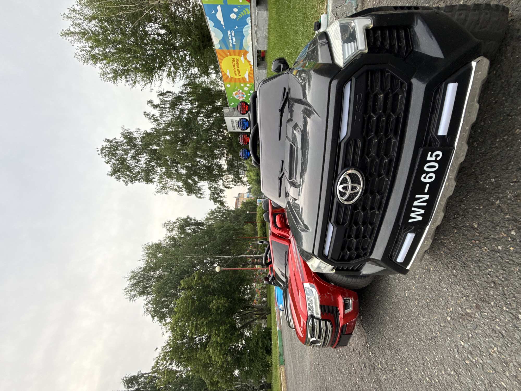 In Krasnoyarsk, Russia, on a cloudy day, two electric toy vehicles are parked on a paved surface. The vehicle in the foreground is black and resembles a Toyota Tundra. It features a large grille with a Toyota emblem, headlights, and a license plate reading "WN-605". Above the windshield, three colored lights are mounted. To its left, a red electric toy vehicle, similar in design to a Toyota Land Cruiser, is visible. Behind these vehicles, trees and a colorful mural are present. The mural depicts a yellow sun and blue sky with clouds, along with text in Russian. The overall scene suggests an outdoor recreational area or park.