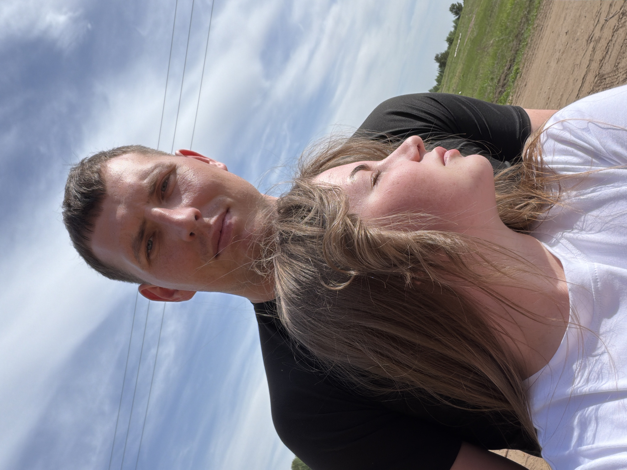 A man and a woman are lying on the ground in a field. The man is wearing a black t-shirt, and the woman is wearing a white t-shirt. The woman is lying on her back with her eyes closed. The man is looking at the camera. The sky is cloudy. There are power lines overhead. The location is near the city of Ural'skoe, Russia. The scene is a close-up shot. The ground appears to be dirt and grass.