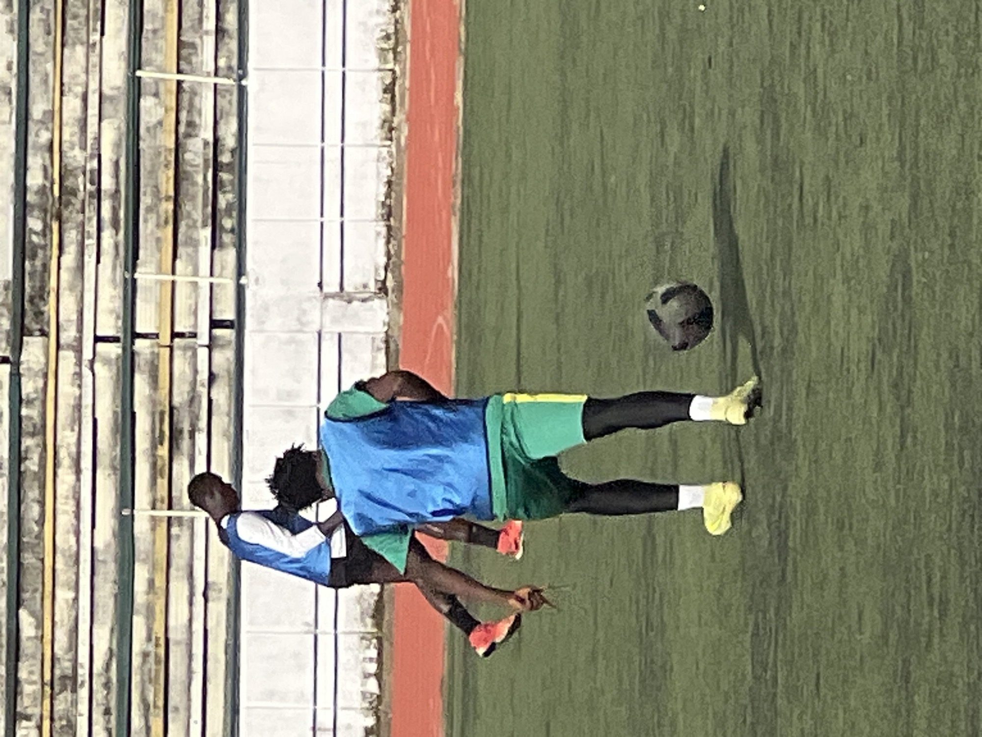 Two male individuals are on a grass field, likely a sports field, in New Stadium, Nigeria. One individual, in the foreground, is wearing a blue vest over a green short-sleeved shirt, green shorts with a yellow stripe, black leggings, white socks, and yellow soccer cleats. This individual is in motion, with their right leg extended and their left leg bent. A soccer ball is in the air to the right of this individual. The second individual, in the background, is wearing a blue and white long-sleeved shirt and black shorts, with orange soccer cleats. This individual is also in motion, with their legs extended as if jumping or kicking. In the background, there are tiered stadium bleachers and an orange running track. The lighting suggests it is daytime.