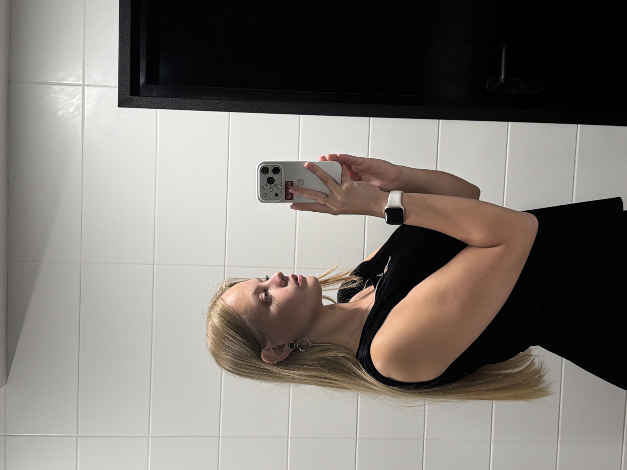 A young woman with long blonde hair is taking a selfie in a bathroom. She is wearing a black sleeveless dress and a white smartwatch. She is also wearing earrings and a necklace. The bathroom has white tiled walls and a black door. There is a black handle on the door. The image was taken in Izhevsk, Russia.