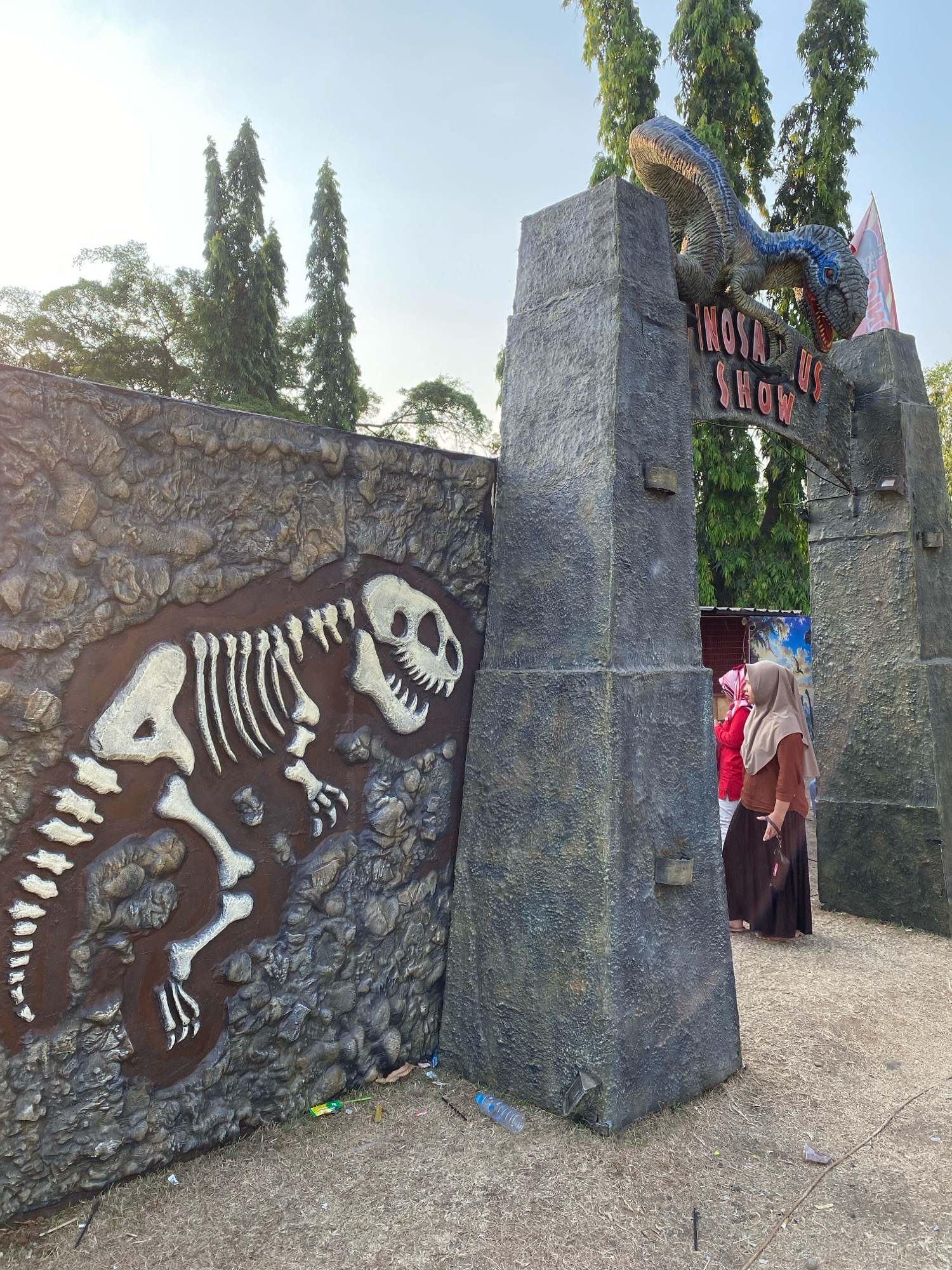The image shows the entrance to a "DINOSAURUS SHOW" attraction, likely in Nyamok, Indonesia, with a large stone archway and a dinosaur sculpture perched on top. To the left of the archway is a wall with a fossilized dinosaur skeleton relief. Two individuals, appearing to be women wearing headscarves, are entering the attraction through the archway. The ground is covered in dry grass and scattered debris, including plastic bottles. Tall trees are visible in the background against a bright sky.