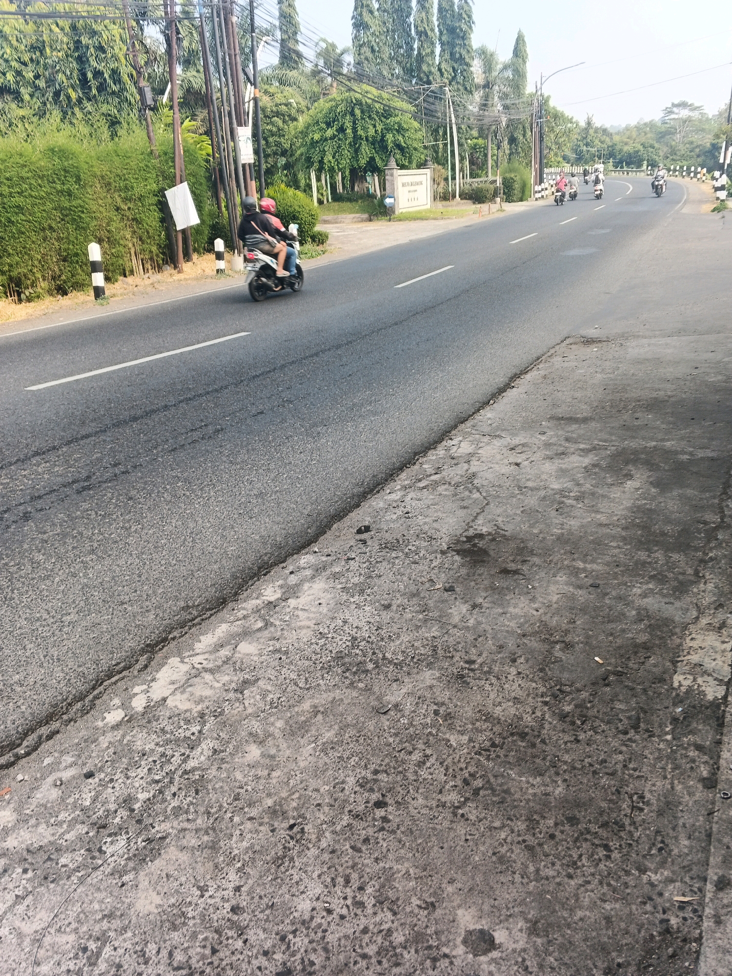 The image shows a road in Ungaran, Indonesia, with a scooter carrying two people in the foreground. The road is asphalt and has a white line marking the lanes. There is a dense green hedge on the left side of the road, with utility poles and wires. A sign for "Melati Balemong" is visible on the left. In the background, there are more trees and buildings. Several other scooters are visible further down the road. The sidewalk in the foreground is made of concrete and appears weathered.