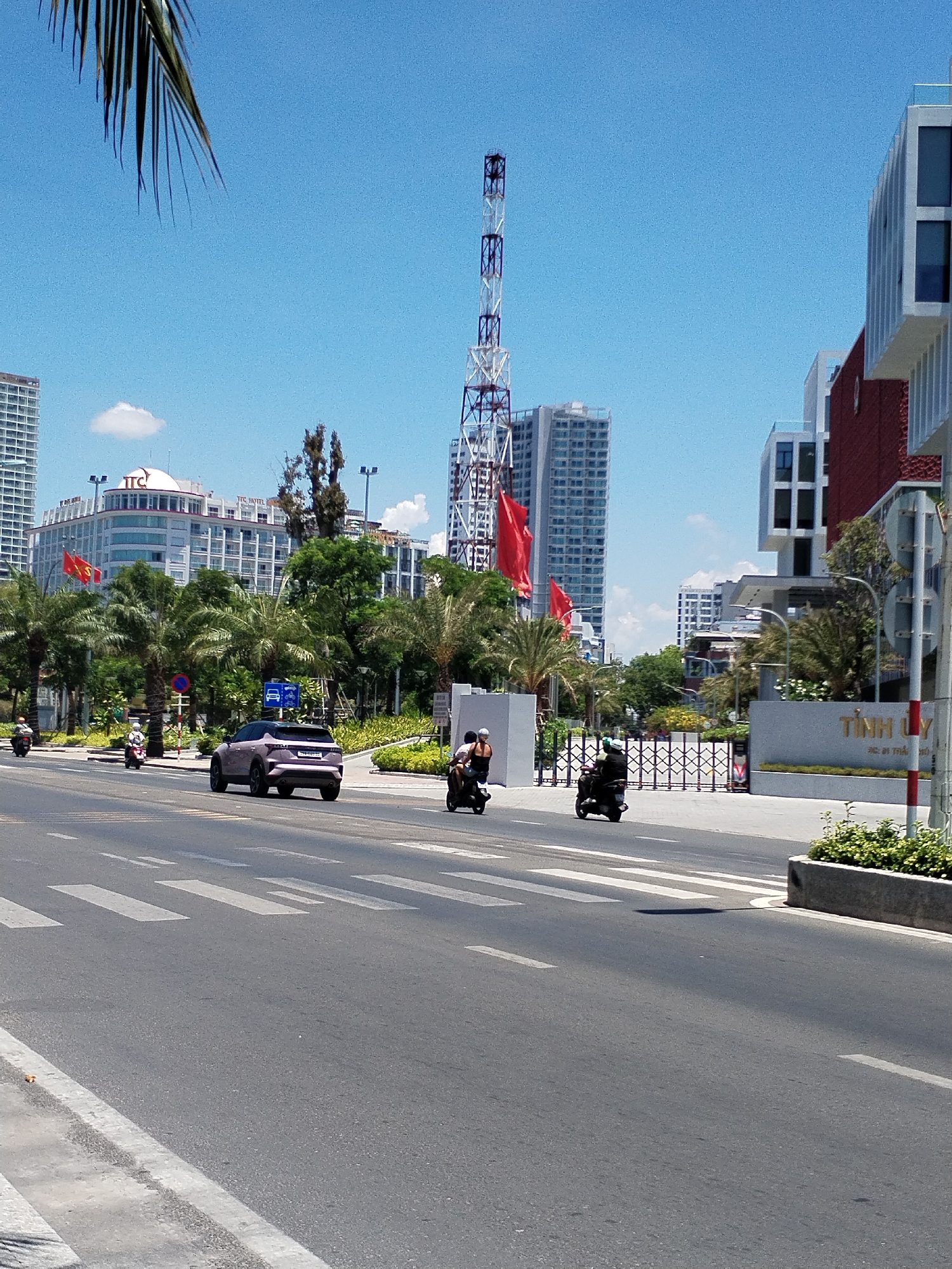 The image depicts a street scene in Nha Trang Ward, Vietnam. A purple SUV is traveling on the road alongside two motorbikes. There are several tall buildings in the background, one with the ITC logo and another with a communications tower atop. There are palm trees lining the road, and Vietnamese flags are flying in front of one of the buildings. There is a pedestrian crosswalk in the foreground. A sign with Vietnamese writing is visible on the right. The sky is blue with some clouds. The overall impression is of a sunny, urban setting.