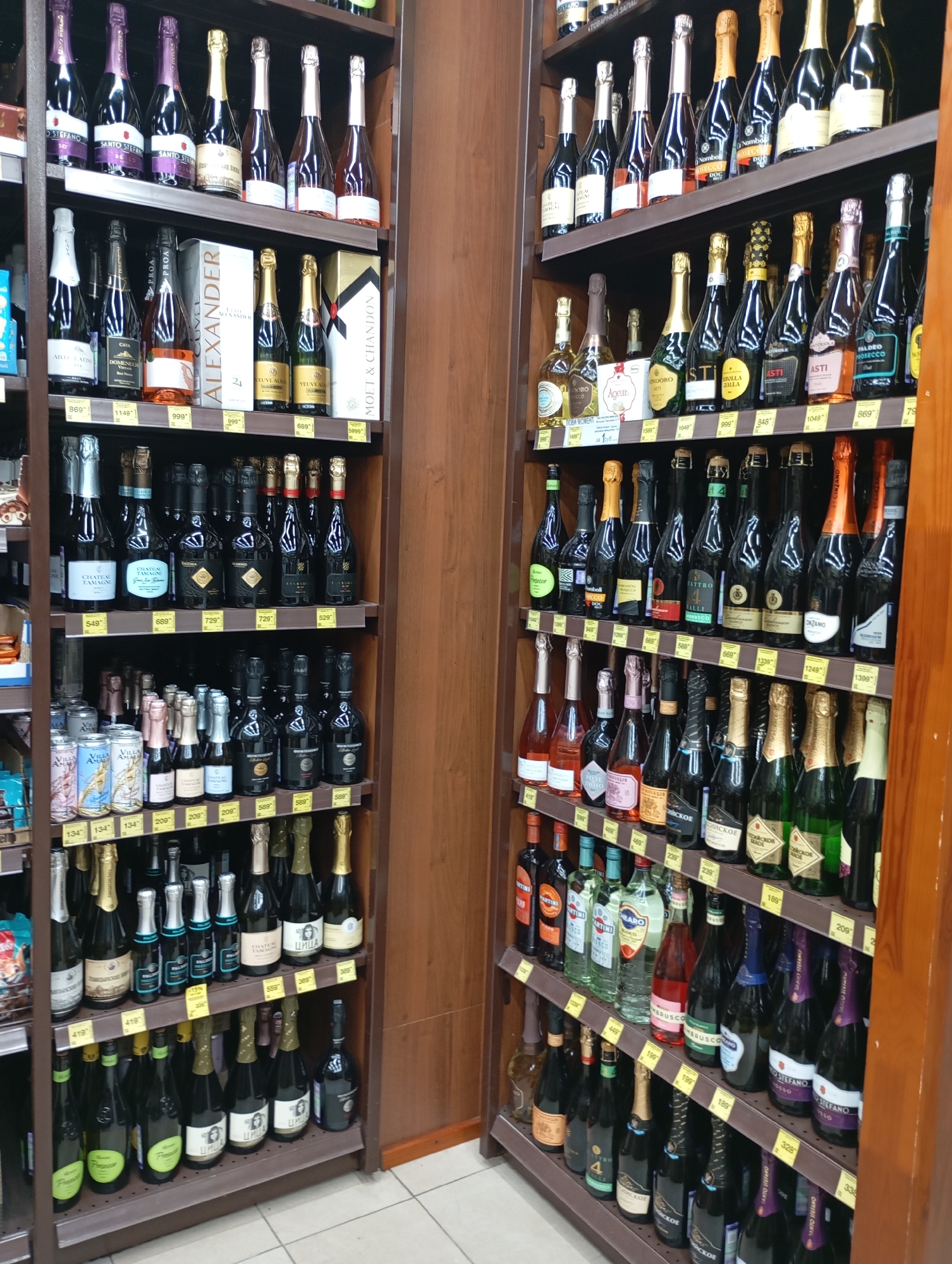This is a photograph of a retail store, likely a wine or liquor store, with shelves stocked with various bottles of alcoholic beverages. The shelves are arranged in vertical rows, with the items displayed from top to bottom. The lighting appears to be artificial, typical of an indoor retail environment. The overall scene depicts a well-organized display of products for sale. The question of Moscow, Russia location is not derivable from the image itself. The image is a close-up shot of shelves stocked with bottles of wine and champagne. Some of the bottles are clear, while others are dark colored. The labels on the bottles vary in color and design, with some featuring text and graphics. The shelves are made of metal and are brown in color. There are also some boxes and cans visible on the shelves, as well as price tags. The background of the image is blurred, suggesting that the focus is on the products displayed on the shelves.