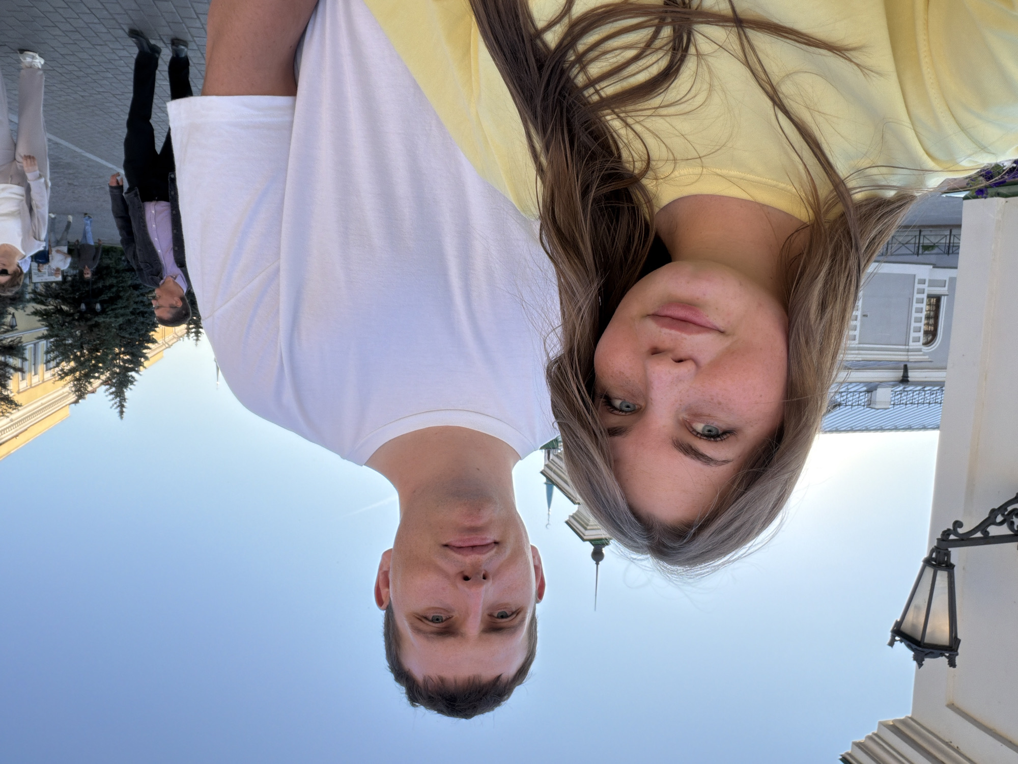 The image features two individuals in the foreground: a woman on the left and a man to her right. The woman has long, light brown hair, light eyes, and light complexion with visible freckles, wearing a plain yellow crew-neck t-shirt. She is positioned slightly forward, looking towards the viewer with a subtle smile. The man has short, dark hair and a light complexion, wearing a plain white crew-neck t-shirt. He is positioned slightly behind and to the right of the woman, looking directly at the viewer with a neutral expression.
The background reveals a clear blue sky. On the far left, a white building facade with a black, ornate wall-mounted lamppost is visible. Behind the subjects, a prominent building with a green roof and a tall, slender spire topped with a crescent moon is discernible, identifying the location as the Kazan Kremlin, specifically near the Kul Sharif Mosque, in Kazan, Russia. Further to the right in the midground, a yellow architectural structure with multiple windows is visible, partially obscured by several coniferous trees. A paved street or pathway occupies the lower right background, where multiple pedestrians are observed, including a man in a dark jacket and trousers walking right, and a woman in a light grey tracksuit walking left. The scene is well-lit by natural sunlight.