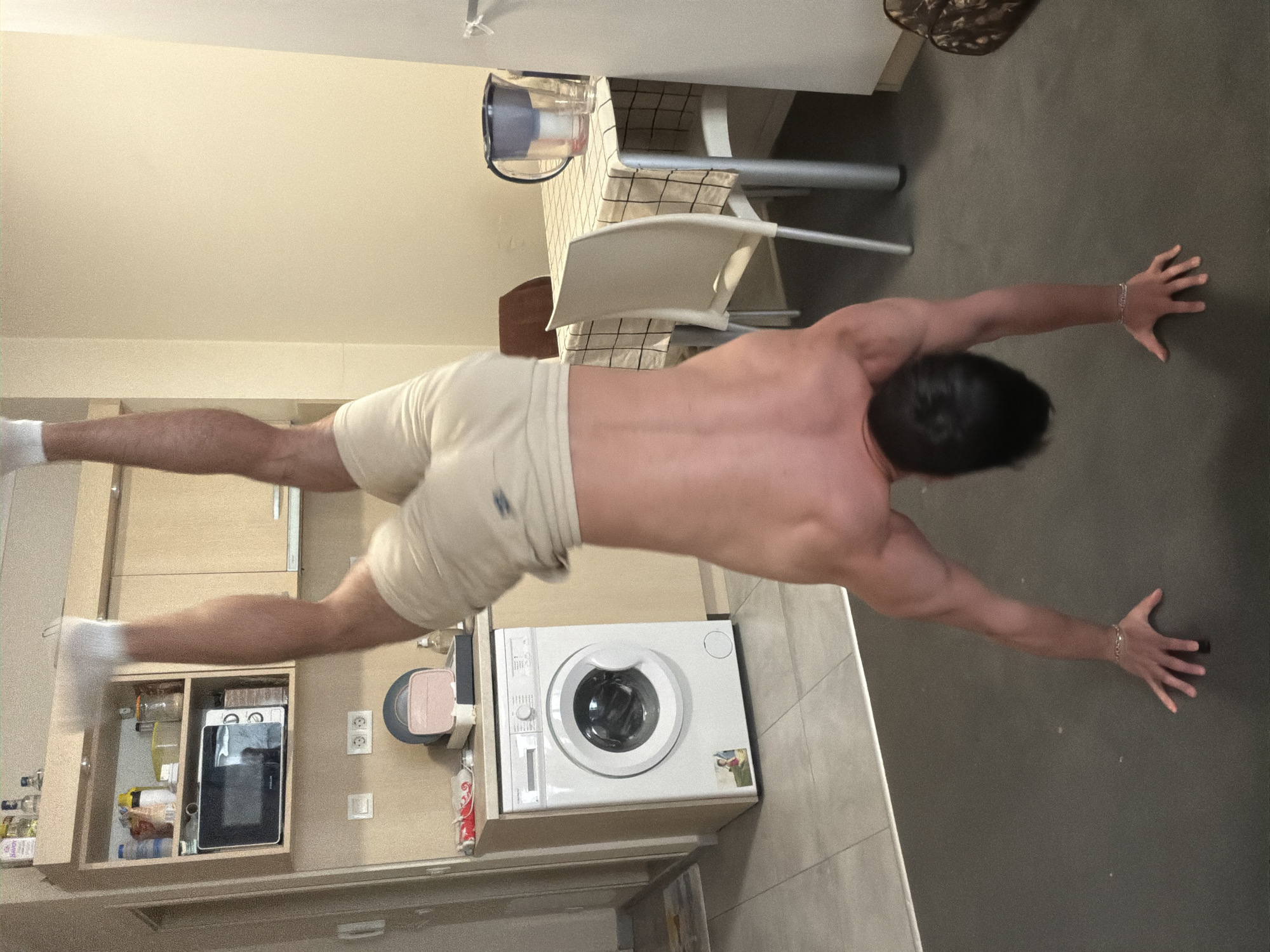 A shirtless man with a muscular physique, wearing light beige shorts and white socks, is performing a "superman" exercise in an indoor setting. He is positioned face down, with his arms fully extended forward, hands flat on the dark gray floor, and his legs stretched backward and raised, parallel to the ground. His head is turned to his right, concealing his face.
The scene appears to be an apartment, combining a kitchen and living/dining area. The flooring is a dark gray material, possibly large tiles or polished concrete, which transitions to lighter tiles near the kitchen cabinets on the left side of the frame. The walls are light beige, and the ceiling is white.
In the background, to the left of the person's body, a kitchen area features light-colored wooden cabinets. An open cabinet reveals a microwave oven and various bottles and jars, one of which has "SAKOR" visible on its label. A white washing machine or dryer is built into the cabinetry. To the right of the person, a dining table with a black and white checkered tablecloth is visible, along with white chairs and a clear pitcher on the table. The lighting is uniform, suggesting artificial indoor illumination. A small, dark, embroidered logo is present on the man's shorts. The overall impression is a domestic environment in Sofia, Bulgaria, where a fitness activity is taking place.