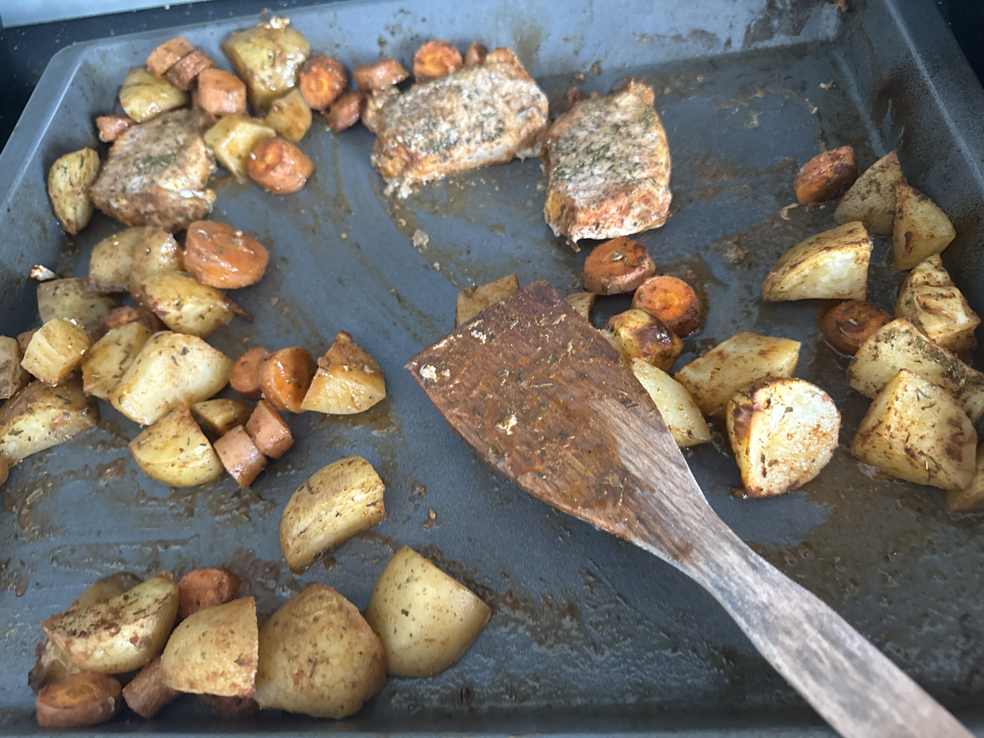 The image depicts a used baking sheet with remnants of a meal. There are scattered pieces of roasted potatoes, carrots, and two seasoned pork chops. A wooden spatula lies on the baking sheet, partially covering some of the roasted vegetables. There is no indication of the location being Sofia, Bulgaria.