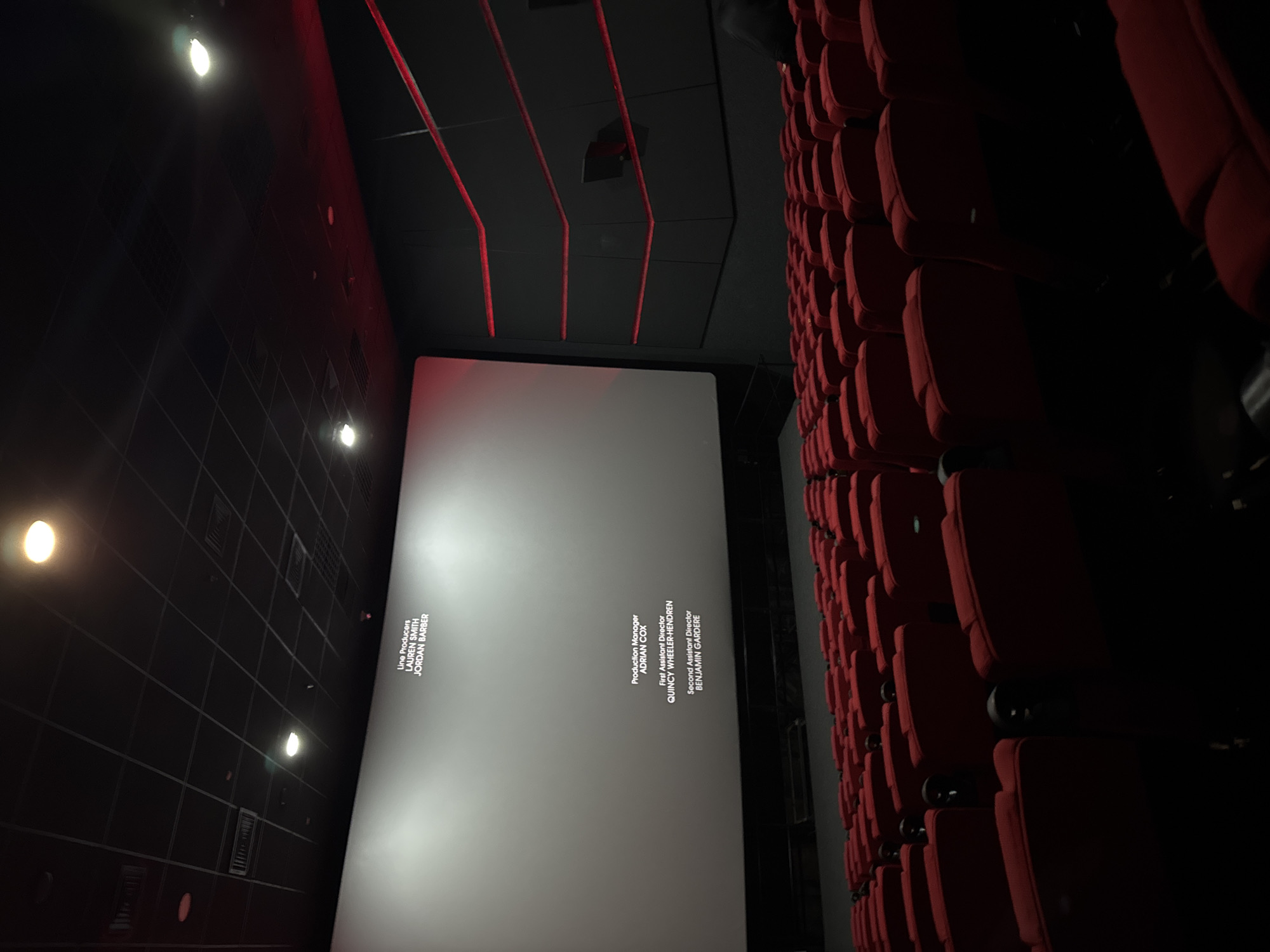 The image shows an empty movie theater with red seats. The screen displays text from the movie credits, including "Line Producers LAUREN SMITH JORDAN BARBER", "Production Manager ADRIAN COX", "First Assistant Director QUINCY WHEELER-HENDREN", and "Second Assistant Director BENJAMIN GARDERE". The lighting is dim, with some lights visible on the ceiling. The theater appears to be modern with dark walls and red accent lighting. No people are visible in the seats.