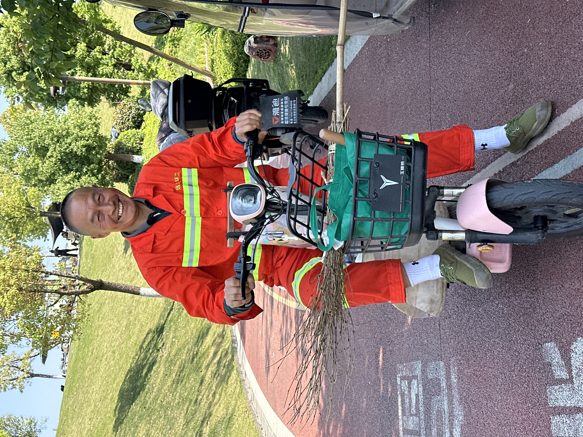 The image shows an adult male riding a scooter on a paved path in what appears to be a park or green space. The man is wearing an orange jumpsuit with reflective stripes, white socks with a logo, and green shoes. He is smiling and looking directly at the camera while holding the scooter's handlebars. A broom is positioned in the scooter's front basket. The scooter is a light color with a pink footrest. The scooter has a black plate with Chinese writing.
The background includes a green lawn, trees, and the edge of a vehicle. The location of the photo is potentially Jiangjin, China based on the information on the black plate that is on the scooter.