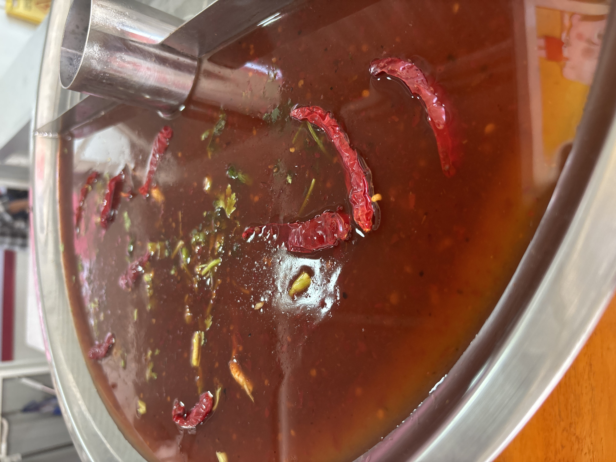 A close-up captures a large container of red-brown sauce, likely a condiment or dipping sauce, at a restaurant or food stall. Dried red chilies and herbs like cilantro are scattered throughout the sauce. The container is made of a shiny, possibly stainless steel, material and has a metal dispensing mechanism attached to one side. The photograph appears to be taken in Baan Bo Phut, Thailand.