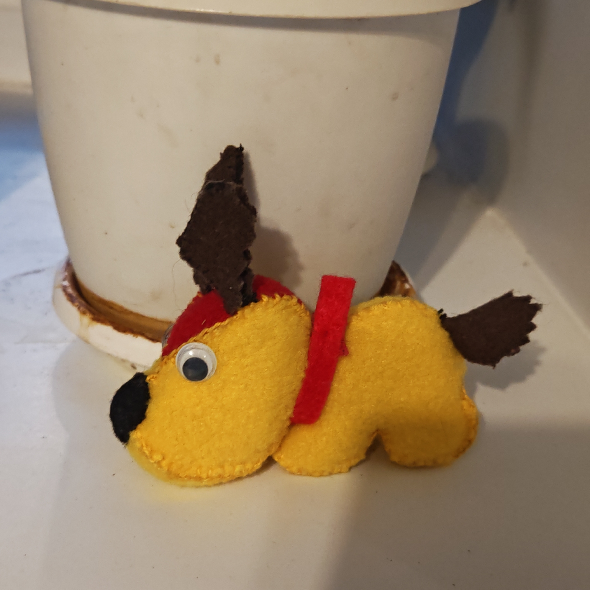 A close-up shot shows a handmade felt dog toy. The toy is primarily yellow, with brown ears and tail, a black nose, a red collar, and a googly eye. It is resting on a white, flat surface. In the background, partially out of focus, is a white flower pot with a saucer. There is no indication of the city of Rostov, Russia.