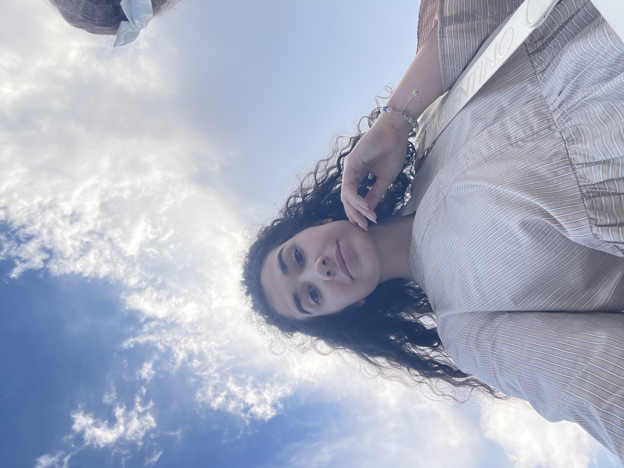 An overhead shot captures a young woman with long, dark, curly hair and light brown skin from the chest up, looking directly into the camera. She is wearing a light brown and white striped dress and a white strap across her chest. Her right hand is raised to her face, touching her chin, and she is wearing a bracelet with colorful beads on her wrist. The background is a bright blue sky with scattered white clouds. The image appears to be taken outdoors, possibly on a sunny day. The location is not specified as Sofia, Bulgaria.