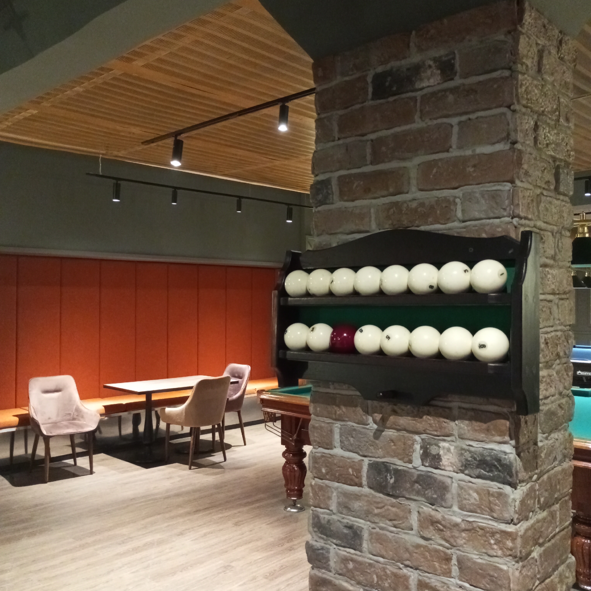The image depicts an indoor billiards hall in Izhevsk, Russia, likely during the evening or night, given the artificial lighting. The scene is set in a modern establishment with a rustic industrial design.
The primary subjects are the billiard balls displayed on a dark wooden rack mounted on a brick pillar. There are two shelves of balls, with the top shelf holding ten white balls, some marked with numbers. The bottom shelf also contains white balls, but with a distinct maroon-colored ball in the center. To the right of the pillar, a portion of a billiards table with a green felt surface is visible.
In the foreground and midground, the setting includes a seating area. Two upholstered chairs, possibly for waiting or resting, are placed on a light wooden floor. Behind these chairs, a long, orange upholstered bench runs along an orange wall, suggesting a comfortable lounge area. A white rectangular table stands between the chairs and the bench.
The architectural details of the establishment are evident, including the textured brick pillar and a slatted wooden ceiling with recessed track lighting. The overall ambiance appears to be calm and relaxed, typical of a leisure venue. No people are visible in the image. There is no discernible text in the image.