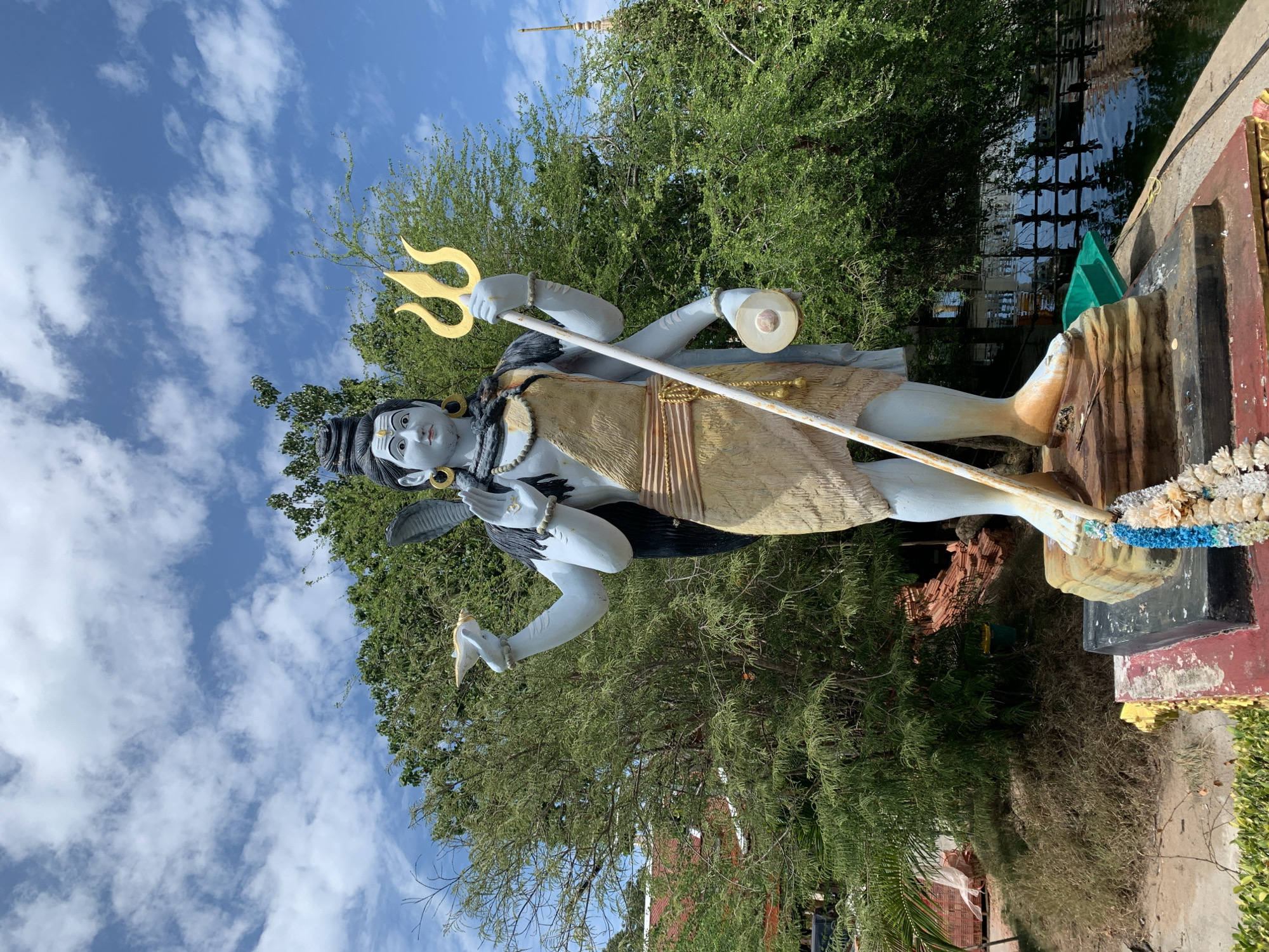 The image shows a full-body statue of the Hindu deity Shiva in front of lush green trees and a blue sky with white clouds. The statue is light blue with several arms, holding various objects including a trident. Shiva is adorned with traditional Indian jewelry and clothing. The statue stands on a tiered platform, which is decorated with flowers. In the background, there are more trees, a body of water, and a glimpse of a building. The overall scene appears to be outdoors, possibly in a temple or park setting, and is illuminated by natural daylight.