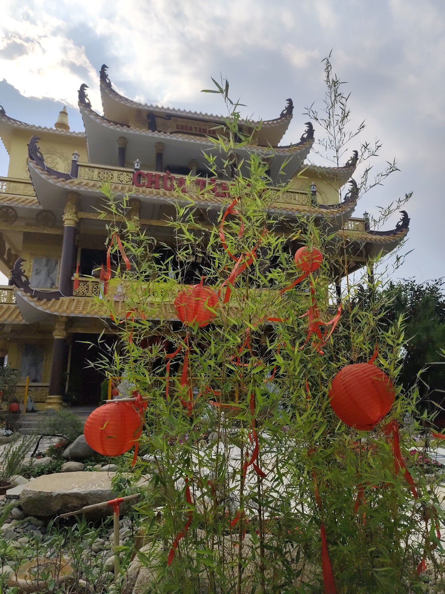 The image captures a scene in Xuân Hương - Đà Lạt, Vietnam, featuring a multi-tiered, yellow Buddhist temple as the backdrop. In the foreground, a cluster of green bamboo shoots is adorned with red ribbons and several spherical red lanterns, suggesting a festive or celebratory context. The ground surrounding the bamboo is covered with small pebbles and rocks, typical of a garden setting. The sky overhead is partly cloudy, indicating daylight. There are no people visible in the frame.