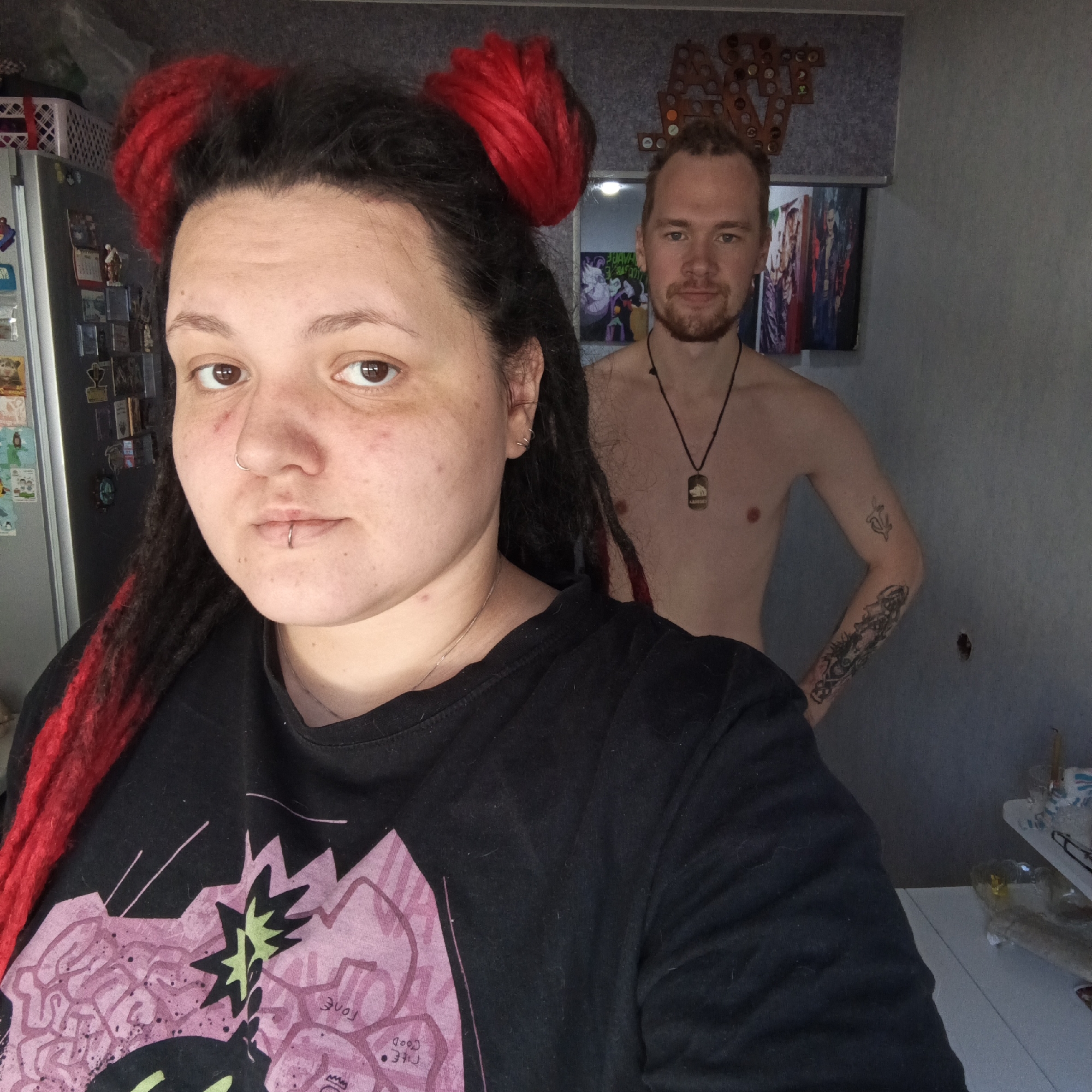 This image depicts two individuals within a residential interior in Chaikovsky, Russia. In the foreground, a female subject with dark dreadlocks featuring bright red sections styled into two buns and wearing a black t-shirt with a pink and green graphic is visible from the chest up. She has a silver hoop piercing in her left nostril and a silver labret ring piercing below her lower lip, and is looking directly at the camera.
Positioned behind her and to the right, a bare-chested male subject with light brown hair and a short beard is visible, also looking directly forward. He wears a black cord necklace with a rectangular pendant featuring an animal emblem. Tattoos are present on his right arm, including a smaller, indistinct design on the bicep and a larger, intricate pattern covering his right forearm.
The background shows a light grey wall. To the left, a stainless steel refrigerator covered with magnets and small items is visible, topped by a white woven plastic basket. To the right, above the male subject, a decorative wooden or similar material sign with circular insets is mounted. Below this, a flat-screen television displays colorful imagery. A white counter or table with various transparent containers, glassware, and packaging is present on the far right. A small circular hole is visible in the grey wall to the right of the male subject's torso.