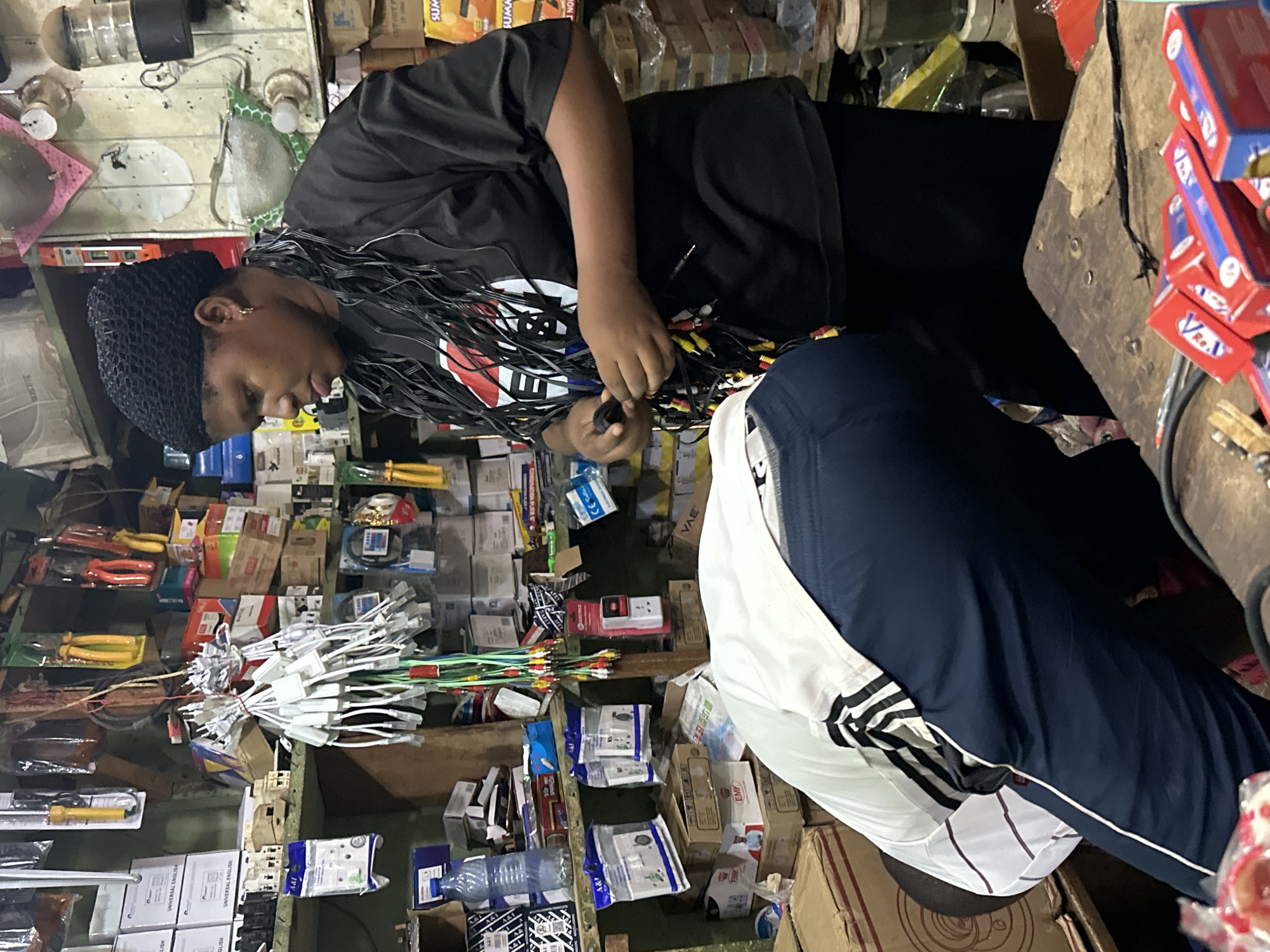The image depicts an indoor scene, likely a small shop or market stall, filled with various electronic accessories and hardware. In the foreground, a person wearing dark blue pants and a white shirt with black stripes is bent over, partially obscured. Standing behind them, a woman wearing a black head covering and a black t-shirt is holding a bundle of multi-colored cables. The shelves behind them are packed with boxes, cables, tools like pliers, and electrical components. The overall impression is of a retail environment stocked with electrical goods. The image appears to have been taken in Barden Yana, Nigeria.