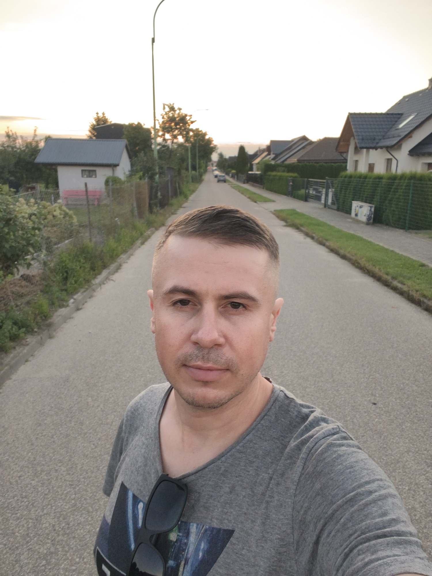 The image is a selfie taken by a Caucasian male with short, dark hair and a light complexion. He is wearing a grey t-shirt with a graphic print on the front and has sunglasses clipped to the collar. The man is standing on an asphalt road in a suburban area, likely in the city of Tczew, Poland. The road is lined with houses, hedges, and trees, suggesting a residential neighborhood. The sky is overcast and light, indicating it might be early morning or late evening. The overall impression is a casual, everyday scene in a quiet Polish town.