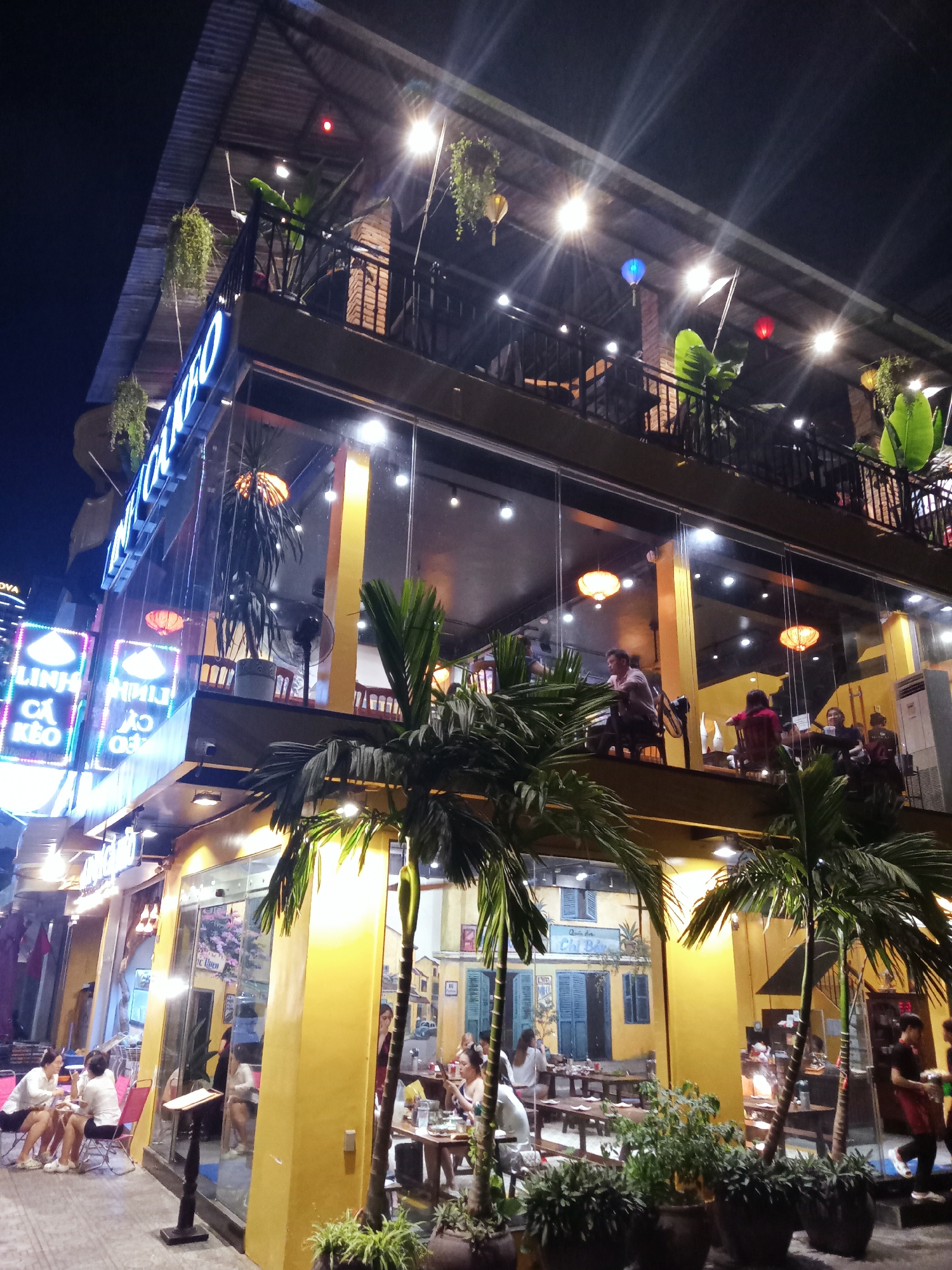 An eye-level shot captures a vibrant, multi-story restaurant at night in an urban setting, likely Nha Trang, Vietnam, as indicated by visible text. The building features a distinctive golden-yellow lower facade with large glass windows, while the upper floors have dark railings and an open-air balcony. Numerous people are visible, dining and socializing across various levels: some sit outdoors at tables on the sidewalk, others inside the brightly lit ground floor, and more are seen on the upper balcony.
The scene is illuminated by an abundance of artificial light sources, including numerous spotlights, hanging lanterns (orange, blue, and red), and prominent neon signs that cause noticeable lens flares. Lush greenery, such as potted palm trees on the sidewalk and hanging plants from the balconies, adds a tropical touch.
Visible text on a neon sign to the left reads "LINH CÁ KÈO" and "TRỘN - NHA TRANG," confirming the location. Another sign on the yellow wall mentions "Quán Ăn Chị Ba" (Sister Ba Restaurant). People appear relaxed, enjoying their meals and conversations in the lively evening atmosphere. The tiled, dark roof of the building is also visible against the dark night sky.