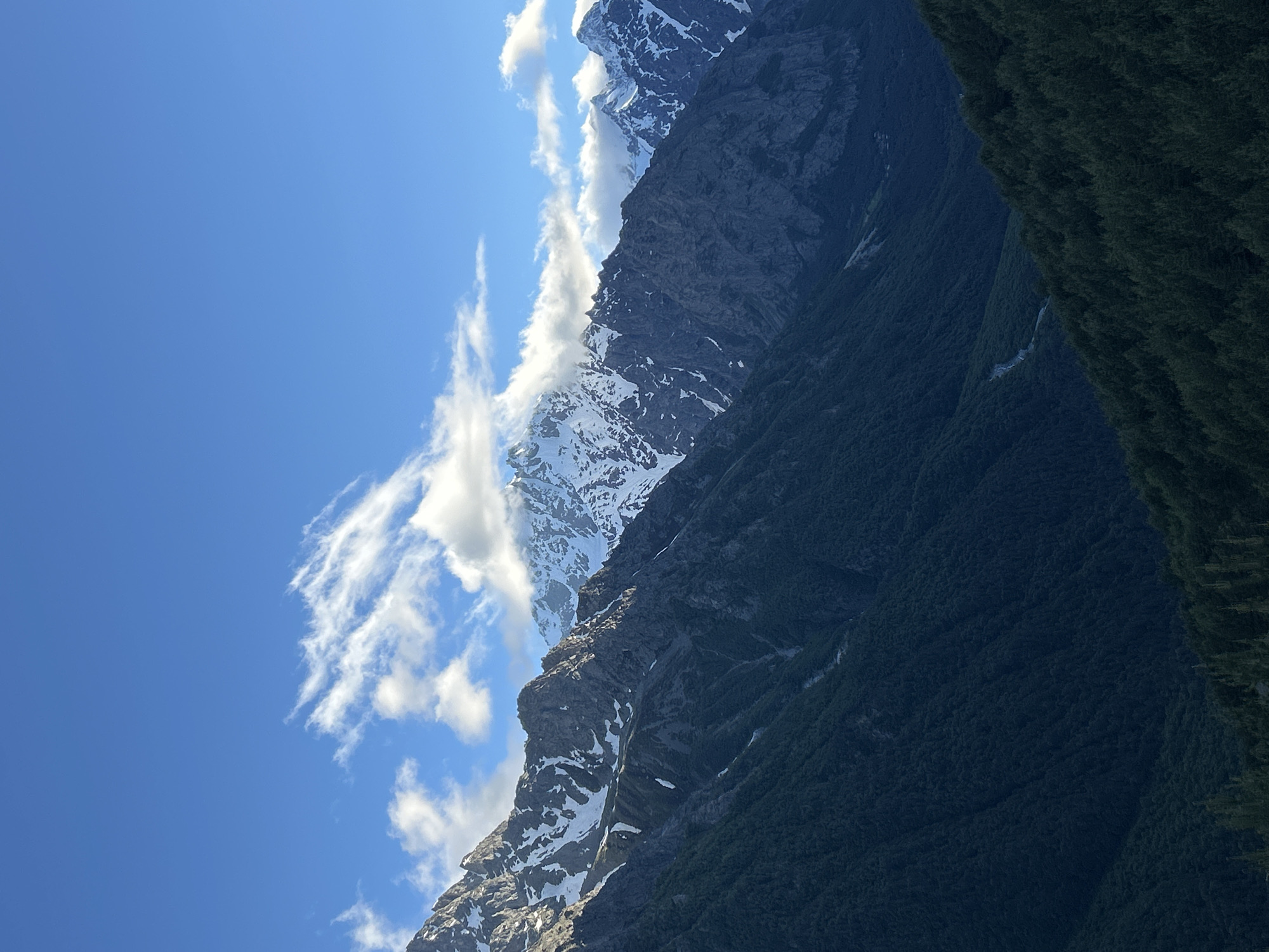 The image depicts a majestic mountain landscape under a clear blue sky. The mountains are rugged and imposing, with rocky outcrops and patches of snow visible on their upper slopes. Wispy clouds drift across the sky, adding a sense of depth and movement to the scene. The lower slopes of the mountains are covered in dense, dark green vegetation, likely a forest. A small, faint stream or river can be seen winding its way down the mountainside through the foliage. The lighting suggests it is daytime, with the sun casting shadows that highlight the contours of the terrain. There are no people or man-made objects visible in the frame, focusing entirely on the natural grandeur of the environment. The overall impression is one of vastness, solitude, and the raw beauty of nature.