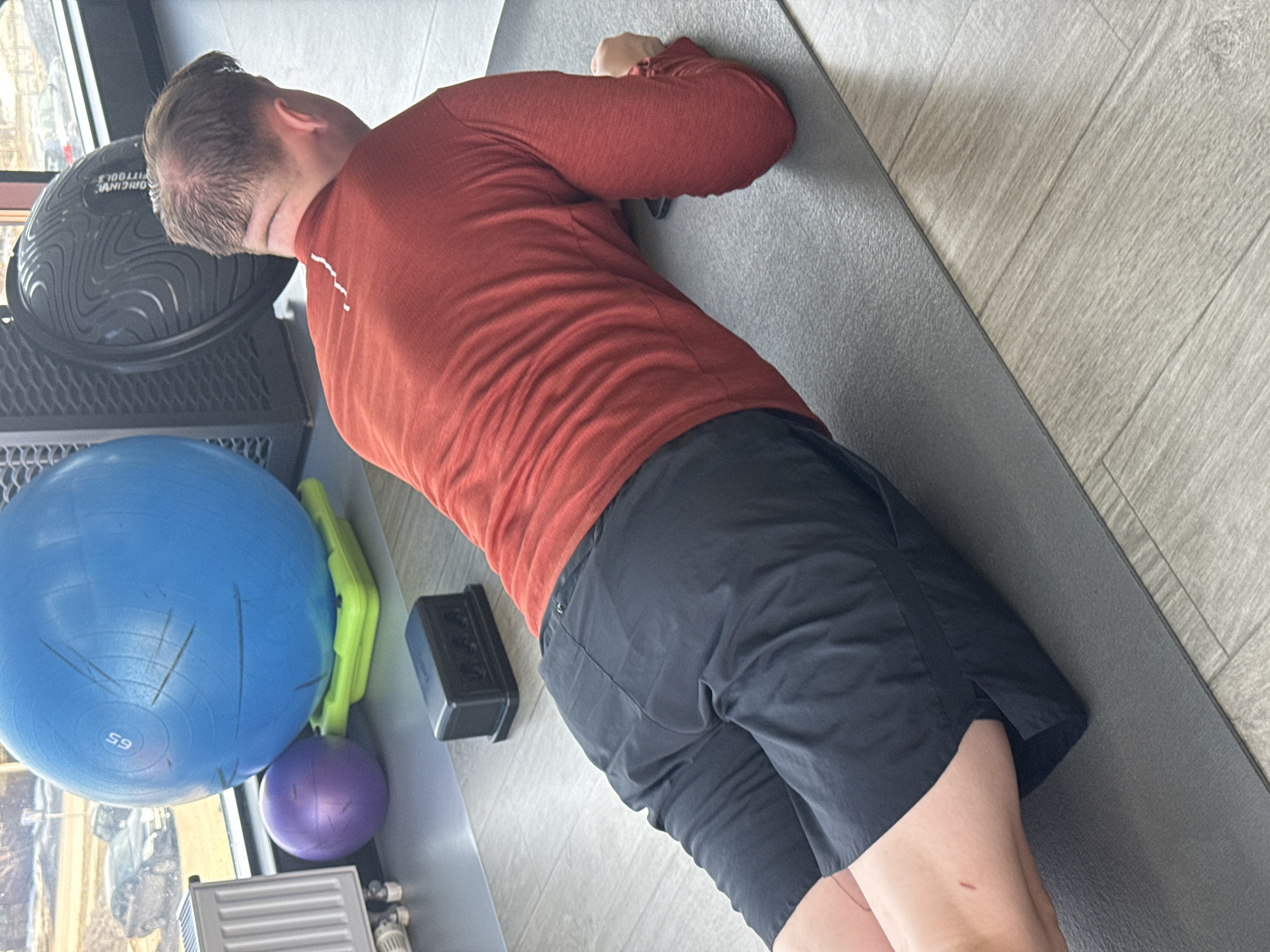 A man is lying on his side on a gray exercise mat in what appears to be a fitness or home gym. He is wearing a long-sleeved red shirt and black shorts. His head is near a black BOSU ball. A blue exercise ball, a green exercise aid, and a purple exercise ball are nearby. The flooring is light wood or faux-wood planks. The location in Votkinsk, Russia, is not discernible from the image.