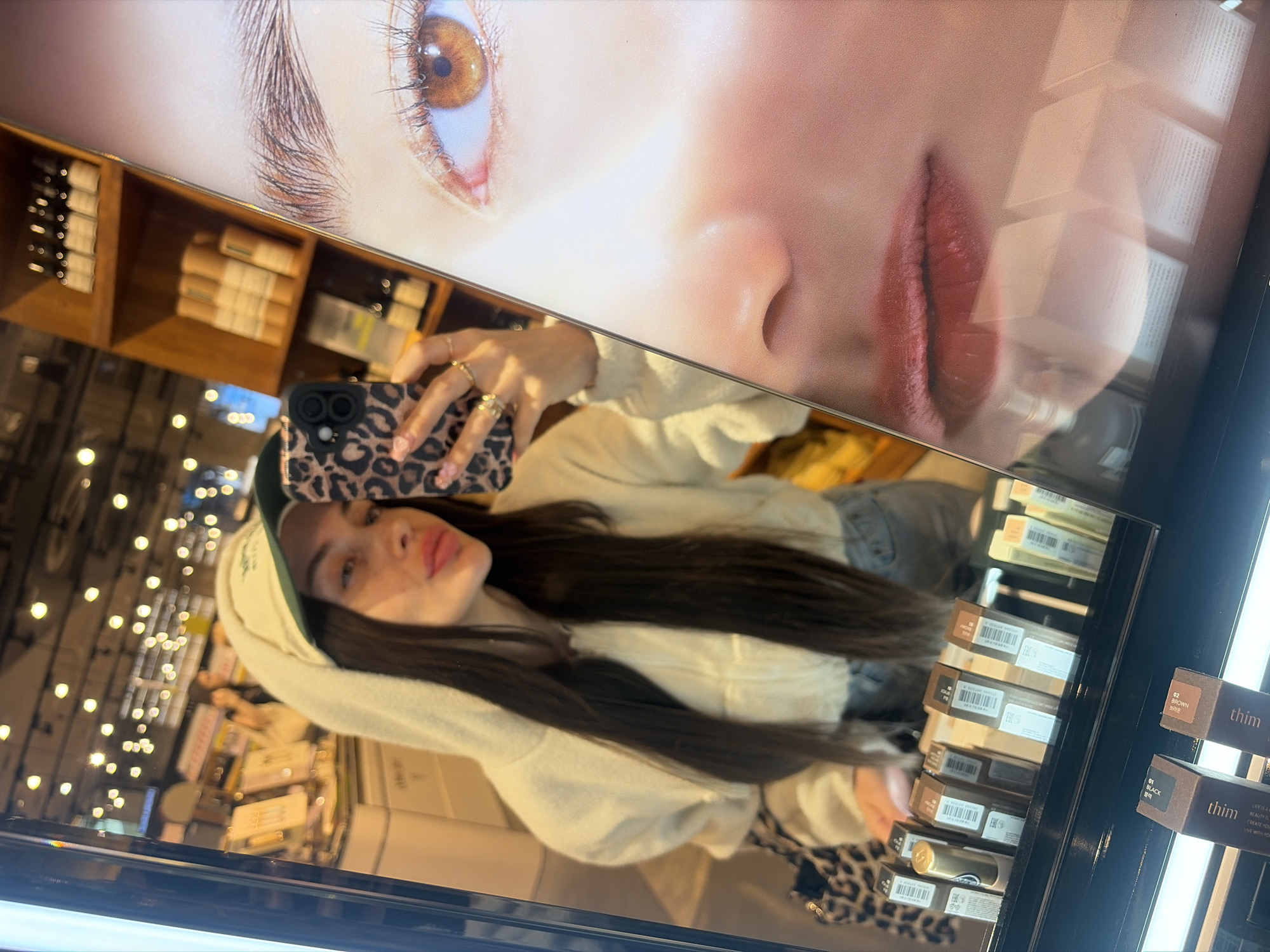 The image is a first-person perspective selfie taken in a retail environment, likely a cosmetics store. A young woman with long dark hair, wearing a white hooded jacket, a cream-colored baseball cap, and light-colored jeans, is holding a smartphone with a leopard print case. She is looking into a mirror that reflects the store's interior. The mirror also shows a large advertisement for makeup, featuring a close-up of a woman's eye, nose, and lips. Products, possibly makeup items, are displayed on shelves in the background and on a counter in the foreground, with visible labels in both English and what appears to be Cyrillic script. The overall lighting suggests an indoor commercial setting. The presence of Cyrillic script and the reference to Saint Petersburg in the prompt indicate that the location is likely Saint Petersburg, Russia.