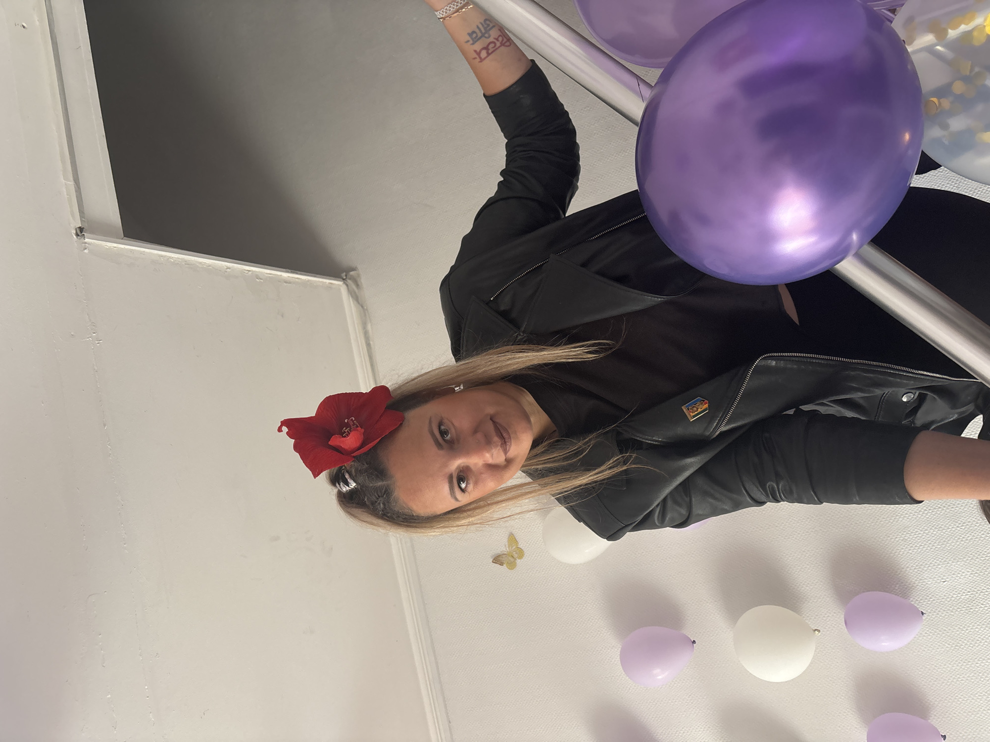 A medium shot depicts a woman indoors, holding a silver railing amidst balloons, her body positioned at an angle, with her feet appearing to be above her head. She has blonde hair and is wearing a black shirt, black jacket, and a red flower in her hair. Visible on her arm is a tattoo. A small butterfly sticker is on the wall, among the scattered purple and white balloons. The scene appears to be in a room with white walls. There is no information available to determine the location as Sochi, Russia.