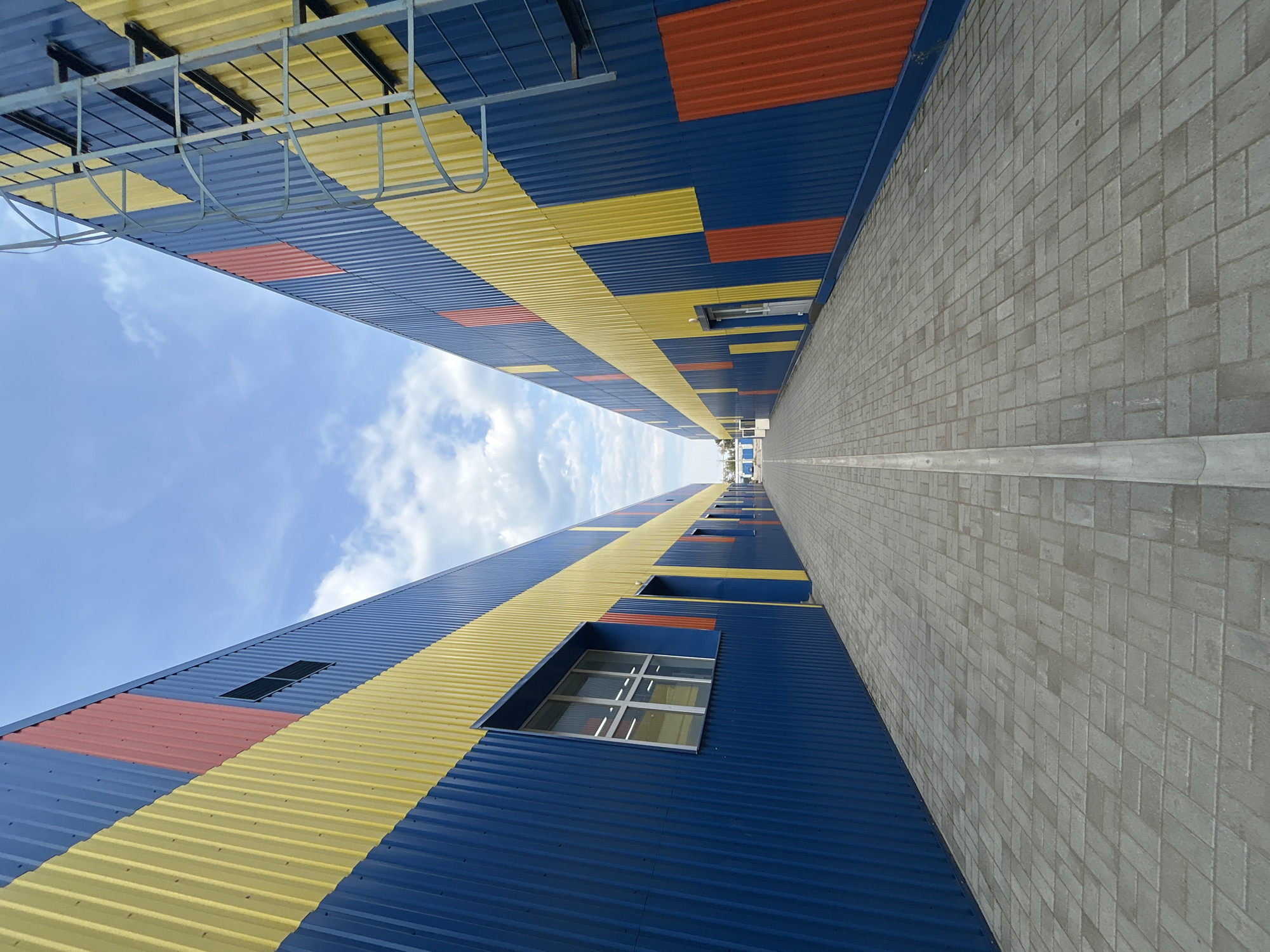 The image depicts a wide, paved walkway between two long, parallel industrial buildings. The buildings have corrugated metal siding in bold, horizontal stripes of blue and yellow, interspersed with rectangular blocks of red. Both buildings feature multiple windows and doors, and one of the buildings has an external metal ladder climbing its side. The walkway is paved with grey rectangular pavers laid in a herringbone pattern, with a concrete drainage channel running down its center. The sky above is blue with scattered white clouds. In the distance, at the end of the walkway, additional structures and a parked vehicle are visible. The scene appears to be in an industrial or commercial area, likely a logistics center or warehouse complex. The location is identified as being in the city of Chaikovsky, Russia. No people are visible in the image.