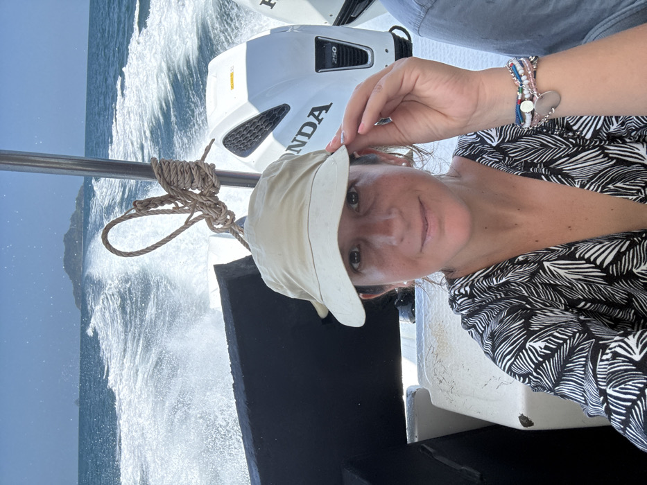 The scene depicts a daytime marine environment from the perspective of a moving boat, identified as near Pak Nam, Thailand. A female subject occupies the central foreground, looking directly at the viewer with a slight smile. She wears a light beige sun hat, a black and white patterned top with a deep V-neck, and multiple beaded bracelets with a silver charm on her left wrist. Her left hand is raised, touching the brim of her hat. To the right, a partial view of another individual's arm, wearing a light blue garment, is visible. In the background, two white Honda 250 horsepower outboard motors are mounted at the boat's stern, generating a substantial white wake across the blue ocean surface. A thick, light-colored rope is coiled and secured to a vertical metal stanchion. On the distant horizon, a small, tree-covered island is present under a clear sky.