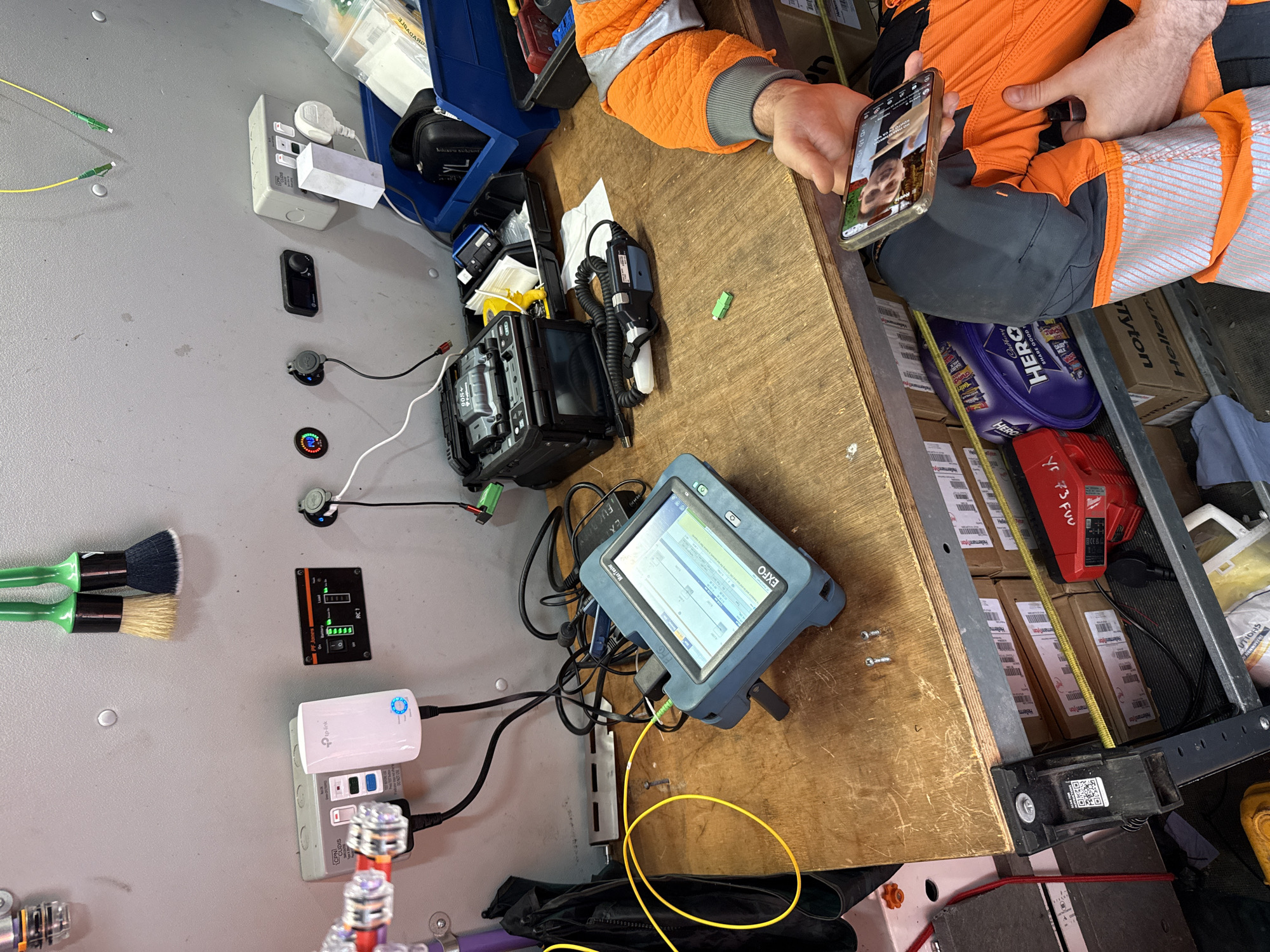 The image shows the interior of what appears to be a work van, with an individual in high-visibility orange and grey workwear on a video call on their smartphone. The van contains various tools and equipment for fiber optic work. A fiber optic fusion splicer and an EXFO MaxTester are prominent on a wooden surface. The van also contains a distribution box, network accessories from TP-Link, brushes, and several boxes, including a Cadbury HERO selection. The environment is cluttered with cables and tools. No specific location details relating to Mid Suffolk, United Kingdom, are visible.