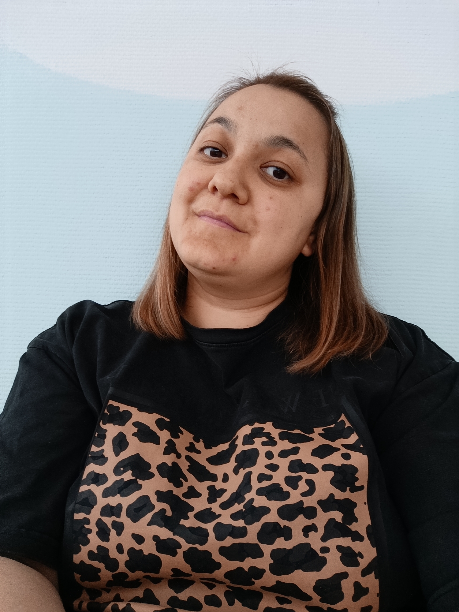 A medium close-up portrait features a woman posing indoors, likely in Chaikovsky, Russia. She gazes directly at the camera with a subtle, closed-mouth smile and a calm expression. Her head is tilted slightly to her right. She has medium-length, light brown hair, parted on her left side, which falls to her shoulders. Her skin is fair, with several small blemishes or marks visible on her forehead, cheeks, and chin. She is dressed in a black long-sleeved top or sweatshirt. The front of her shirt is adorned with a prominent brown and black leopard print pattern, which forms a large 'V' shape towards the neckline. Above this print, on the black fabric, there are faint, dark, illegible letters. The background consists of a light blue wall with a subtle, fine-grained texture, possibly wallpaper or painted. The scene is illuminated by soft, even lighting.