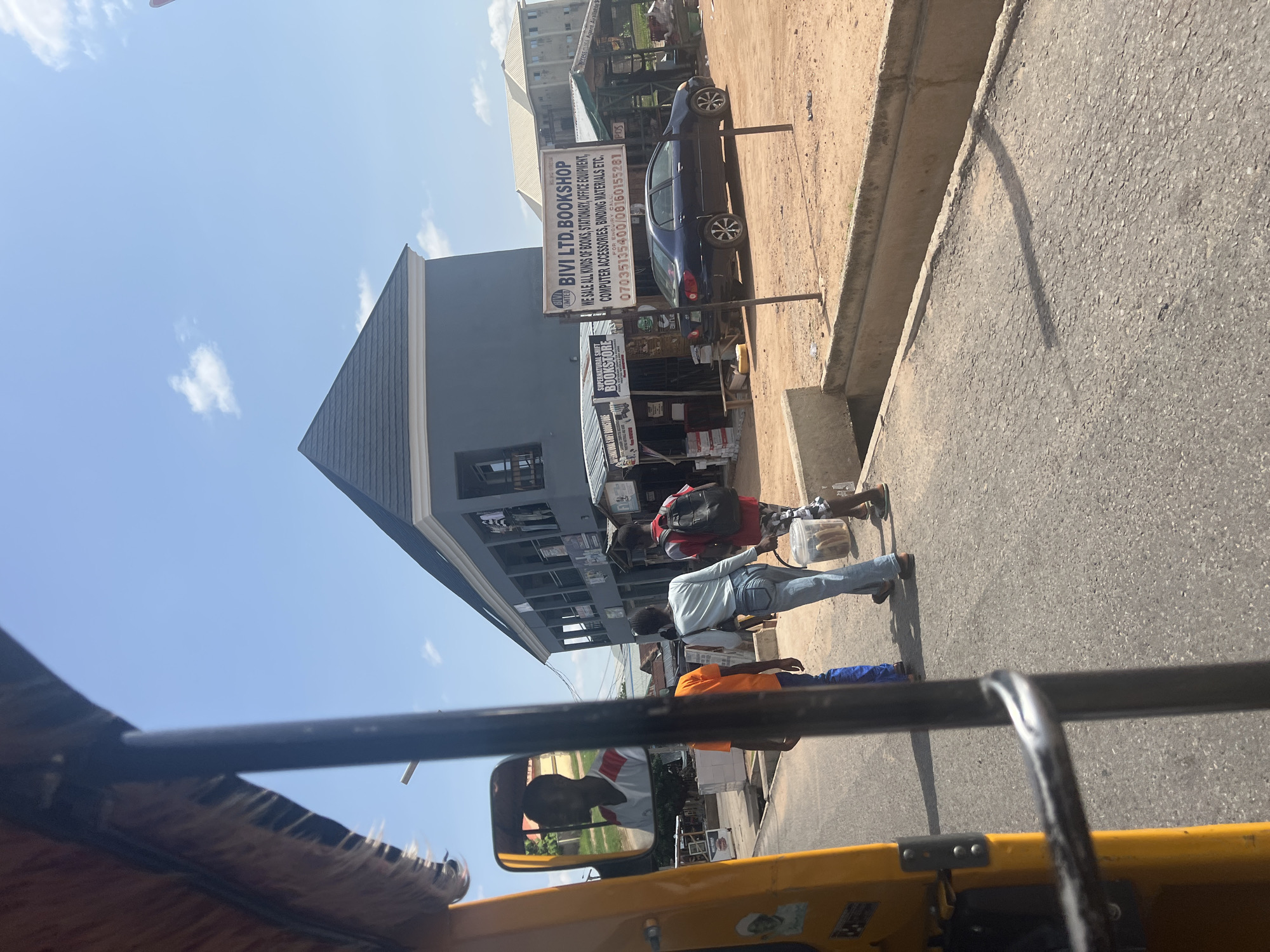 The image shows a street scene in Jalingo, Nigeria, captured from the perspective of a yellow motorized tricycle. In the foreground, the edge of the tricycle's yellow body and a metal bar are visible. A side mirror reflects the face of a man wearing a white shirt with red accents.
On the street, three individuals are walking away from the camera. The person in the center wears a light blue long-sleeved shirt and blue jeans. To their left is a person in an orange t-shirt and blue pants. To the right is a person wearing a red backpack and checkered shorts, carrying a clear plastic container filled with bread.
Several shops and buildings line the right side of the street. A prominent sign reads "BIVI LTD. BOOKSHOP" and lists items sold such as books, stationery, office equipment, and computer accessories. A dark blue sedan is parked in front of the bookshop. Further down the street, other shops and a light-colored building are visible. The environment appears to be a commercial area with a paved road and a concrete drainage channel. The sky is clear blue with a few scattered clouds.