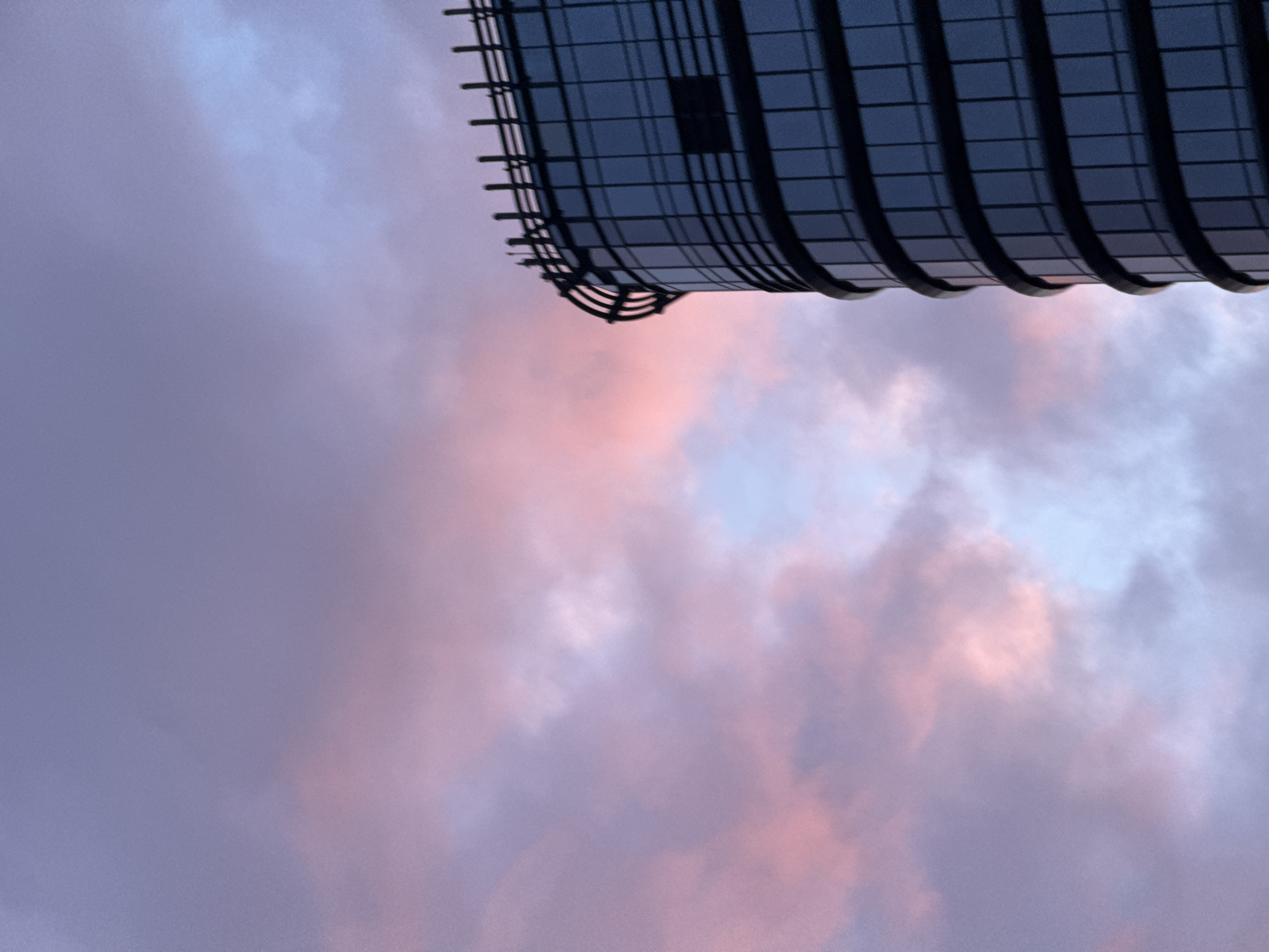 The image presents a modern, curved glass building silhouetted against a vibrant twilight sky in Minsk, Belarus. The building occupies the right portion of the frame, with its dark, reflective facade composed of multiple horizontal and vertical grids forming window panes. A distinctive architectural feature, a curved railing or parapet with several vertical posts or fins, adorns the top edge of the structure. The sky, filling the majority of the left and upper frame, displays a dynamic gradient of colors, transitioning from deep purples in the upper left to soft pinks and oranges near the building, and light blue in patches, indicative of either sunrise or sunset. Scattered, wispy clouds are visible, absorbing the various hues of the sky. No people, discernible actions, or interactions are visible in the scene. The perspective is from a low angle, looking upwards at the urban structure and the atmospheric conditions.