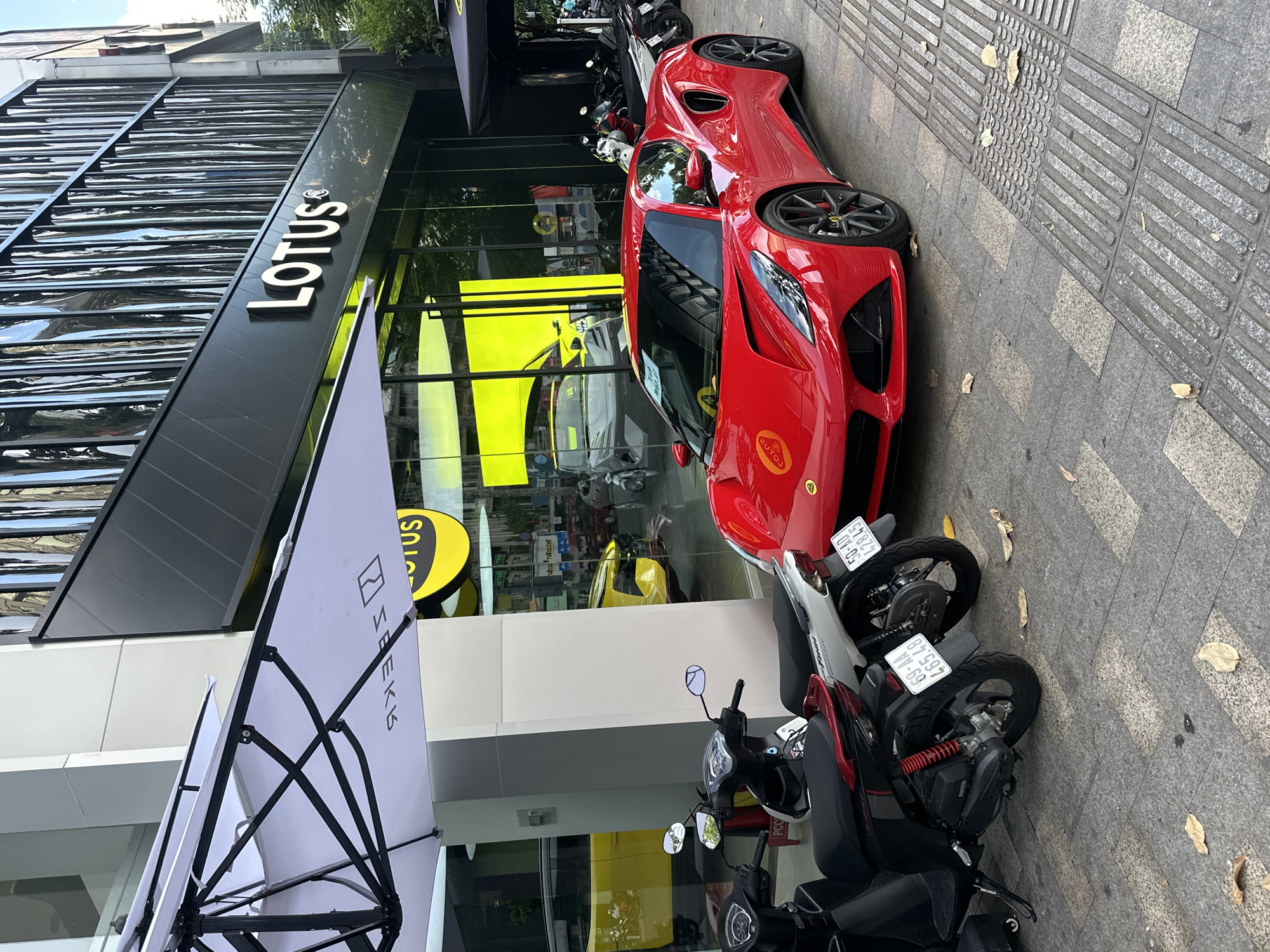 This media file shows a red Lotus sports car parked on a sidewalk in front of a Lotus dealership in Thủ Đức, Vietnam. The dealership has a modern architectural design with large glass windows and a prominent "LOTUS" sign. Several motorcycles are parked on the sidewalk near the car. The scene appears to be set during the daytime, with natural light illuminating the area. The weather seems pleasant, with a few fallen leaves on the ground suggesting an outdoor setting.
Notable details include the distinctive design of the Lotus car, the branding of the dealership, and the presence of multiple motorcycles, which are common in Vietnam. The overall impression is one of an urban environment, possibly a commercial district, where luxury vehicles and everyday transportation coexist. The image is taken from a slightly elevated perspective, looking down at the car and the surrounding area. No people are visible in the frame, emphasizing the focus on the car and the dealership. The image is oriented vertically, and the content is presented from right to left.