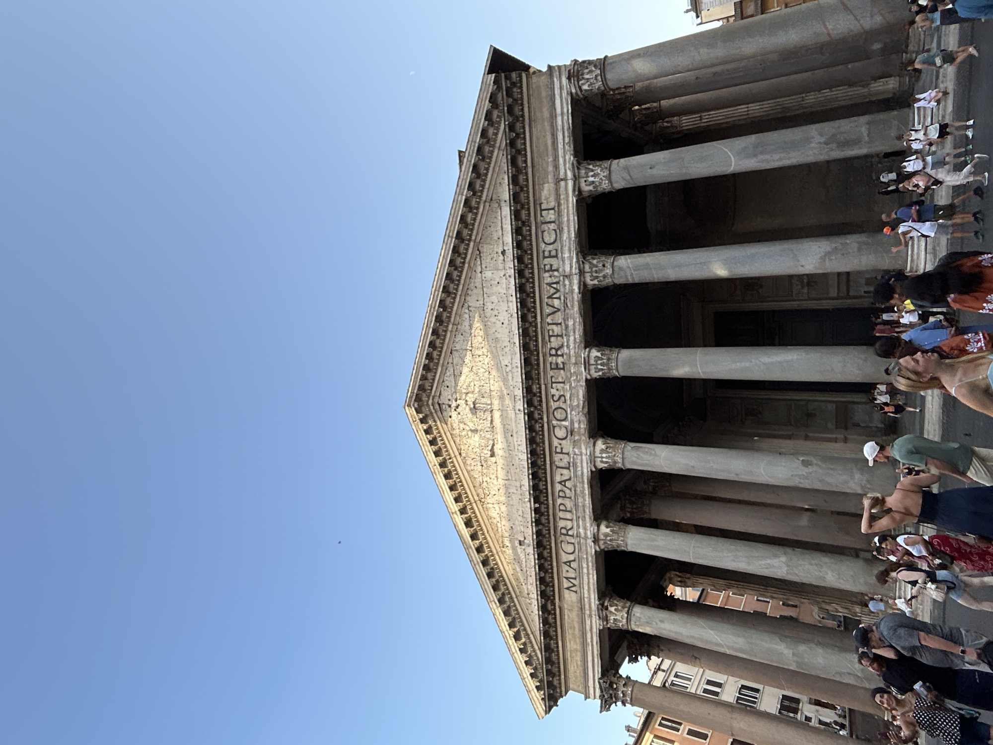 The image shows a low-angle view of the Pantheon in Rome, Italy, under a clear blue sky. The iconic structure, with its massive Corinthian columns and triangular pediment, dominates the frame. The text "M AGRIPPA L F COS TERTIUM FECIT" is inscribed on the entablature, a testament to its history.
A group of tourists are gathered in front of the Pantheon, appearing to be exploring and taking in the sights. They are dressed in casual summer clothing, suggesting a warm, sunny day. Some individuals are taking photos, capturing the grandeur of the ancient building. The scattered presence of people indicates a busy tourist site, typical of a well-known landmark. The bright sunlight and clear sky suggest it is daytime, likely mid-morning or afternoon, and the weather is pleasant. The scene evokes a sense of wonder and appreciation for historical architecture.